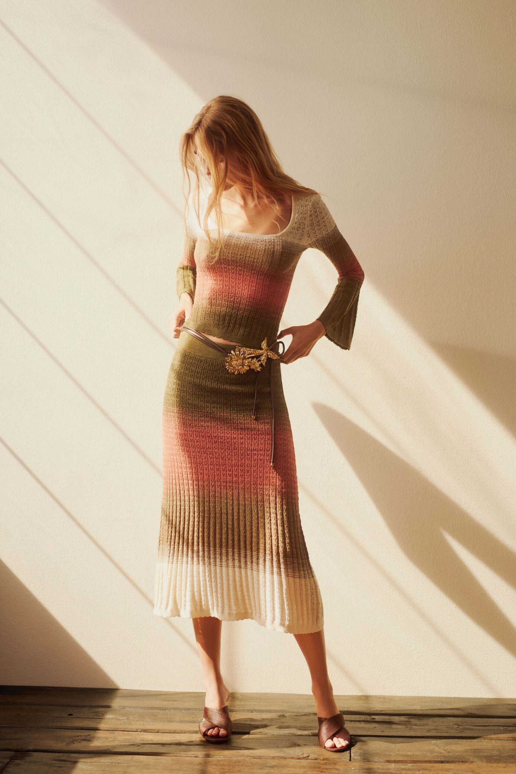 Openwork ribbed knit top, scooped wide and flaring at the cuffs. Matching pointelle knit midi, striped in cream, moss and dusty rose. Thin cord belt knotted through a gilt flower spray buckle. Bronze crossband mules. Late afternoon in a borrowed house, sun bars climbing the wall.
