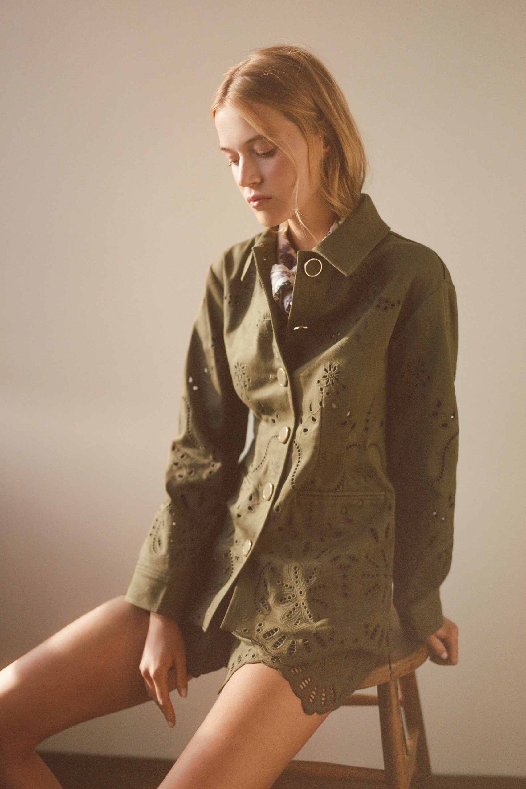 Olive leather jacket punched with floral broderie and big pale horn buttons. Lilac floral neckline peeking out at the open collar. Matching olive leather shorts with a scalloped eyelet hem. Sun-warmed guest room, dressing too early for the garden party.