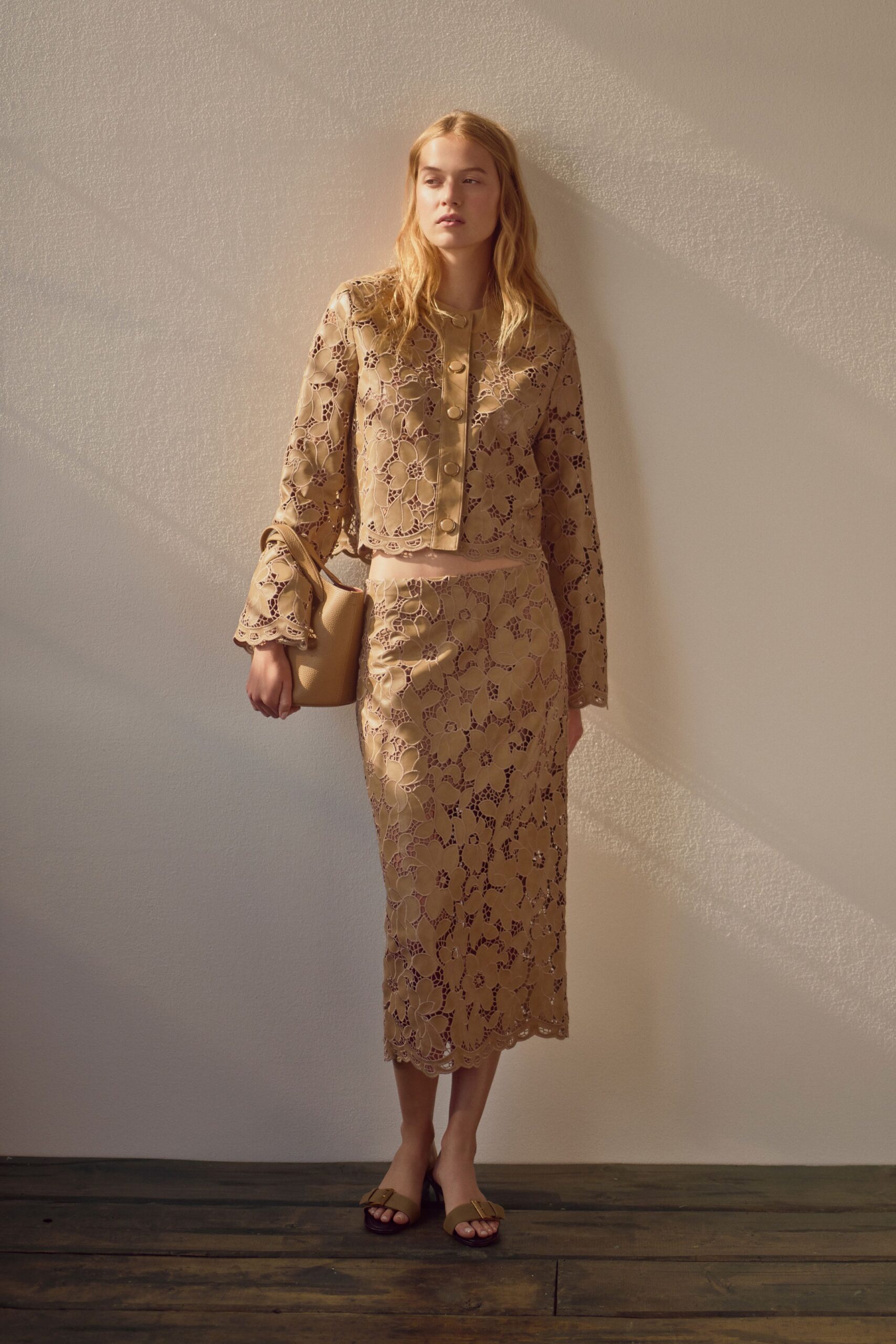 Cropped fawn leather jacket cut in oversized floral guipure, gold buttons down the placket. Matching midi pencil skirt with a scalloped cutwork hem. Small biscuit bucket bag in pebbled leather. Tan slide sandals with a blunt gold buckle. Sun on a stucco hallway before the courthouse wedding starts.