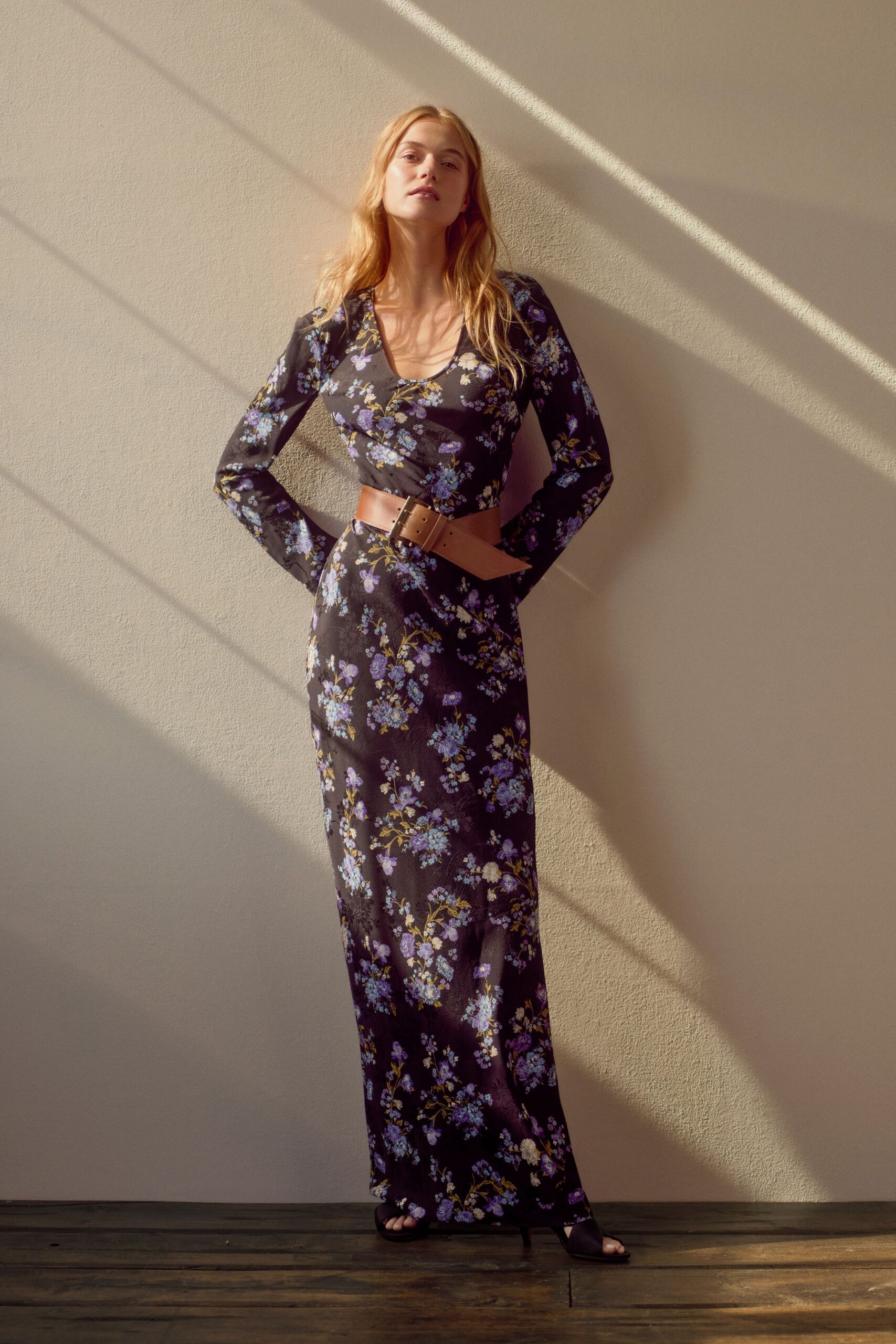 Black satin-column dress scattered with lilac sprigs, scooped low at the chest. Wide tan leather belt, oversized square buckle pulled off-center. Black open-toe mules. Late afternoon in a rented house, sunlight striping the plaster wall.