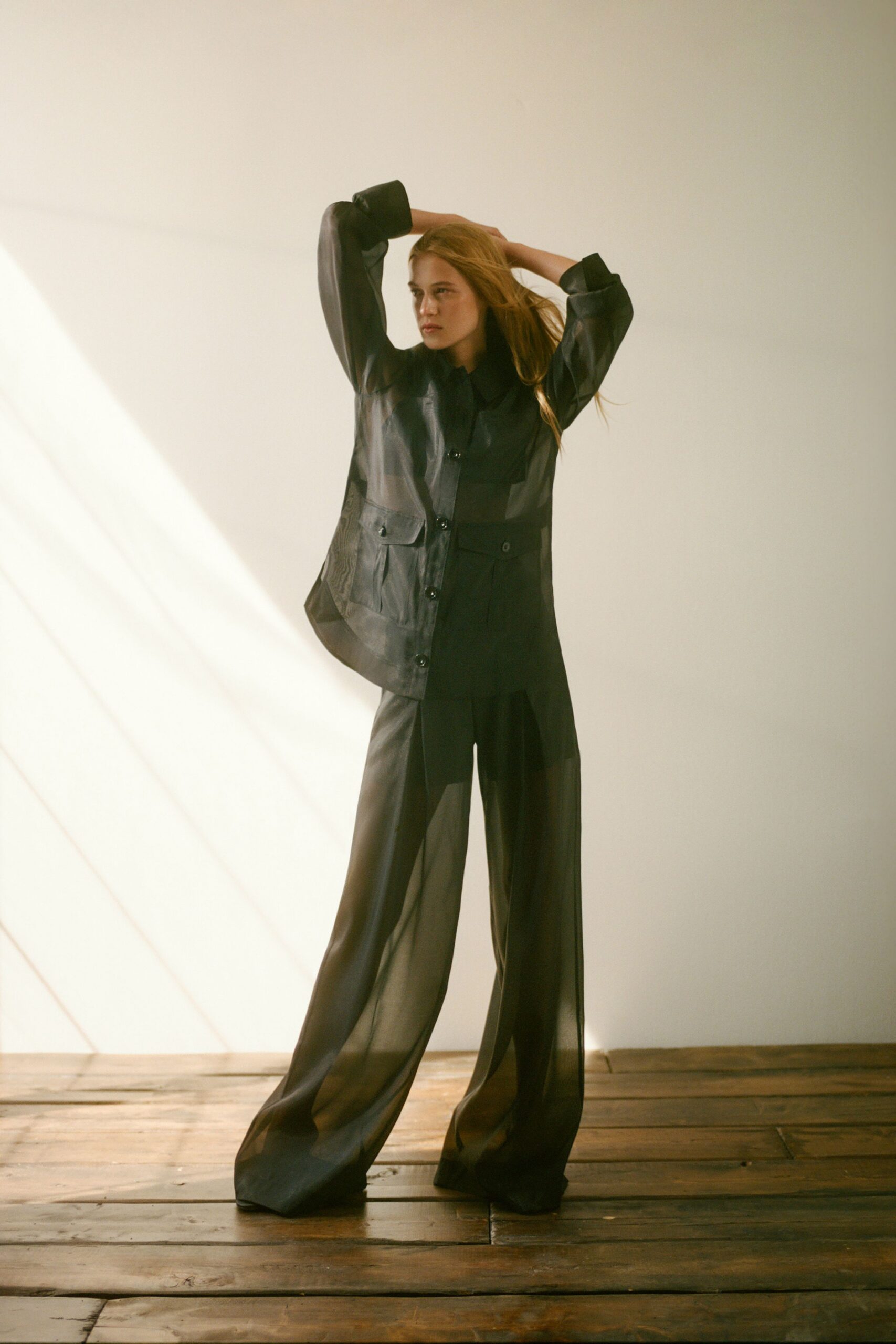 Smoky black organza shirt jacket with flap pockets and glossy buttons. Matching sheer trousers cut extra long, pooling into soft folds. Sun through high windows on a wooden floor before the heat breaks.