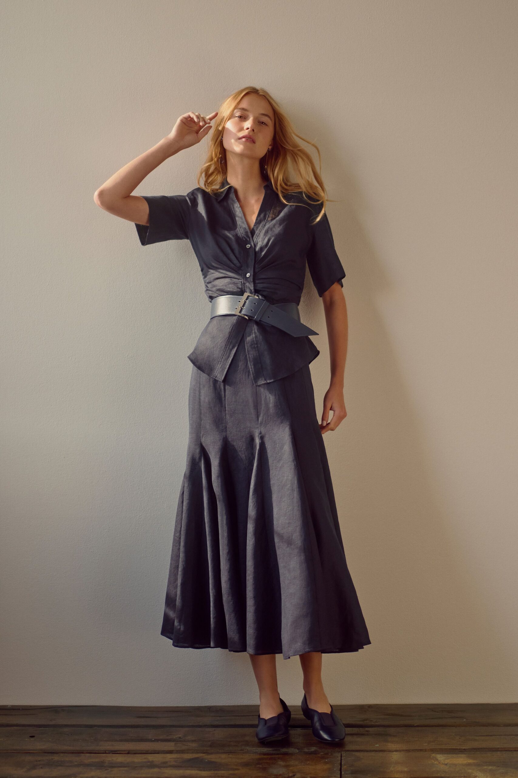 Charcoal sateen blouse with a ruched waist and soft camp collar. Wide matte leather belt, pointed end skewed past the buckle. Matching midi skirt cut in gores that open into a fluid hem. Square-toe black leather flats. Afternoon apartment light, dressed before opening the windows to the heat.