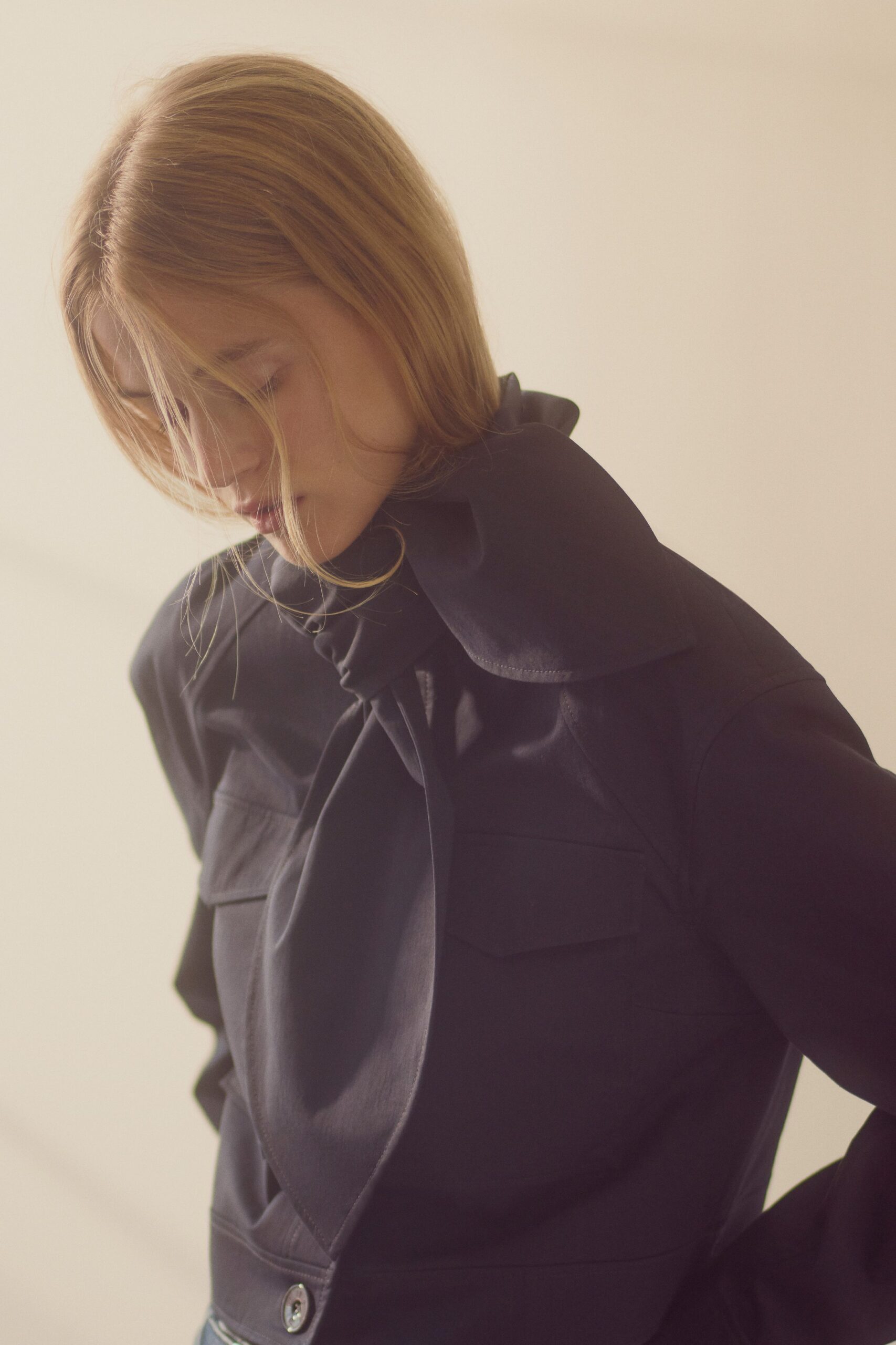 Ink-black blouse with an oversized scarf collar falling in long satin points. Matching cropped jacket, flap pockets and a lone glossy button at the hem. Pale denim just visible below. Morning train after rain, still carrying the smell of someone else's apartment.