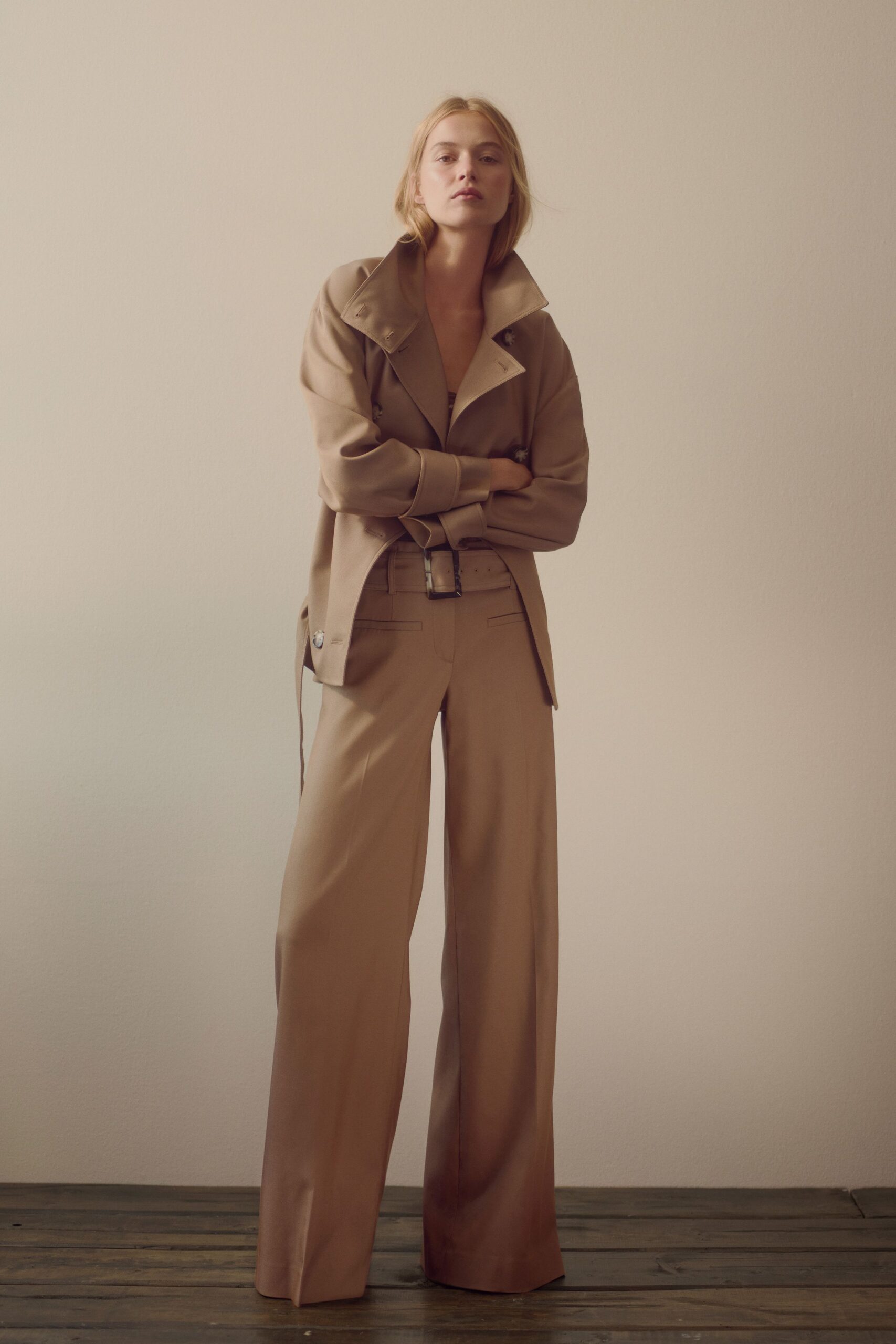 Taupe double-breasted jacket with a popped stand collar and tabbed cuffs. Matching wide-leg trousers, pressed flat with long front creases. Self belt with a square buckle sitting slightly off center. Dark bandeau barely visible at the deep opening. For crossing a silent hotel lobby before the breakfast trays arrive.