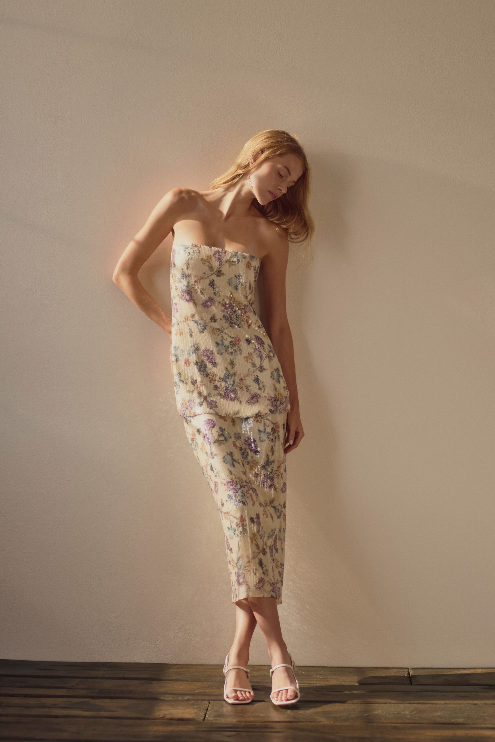 Ivory micro-pleated sequins scattered with lilac and faded blue florals. Matching calf-length skirt, the floral sequins thinning near the hem. Barely-there white ankle-strap sandals. Afternoon wedding light pooling across the rented house floorboards.