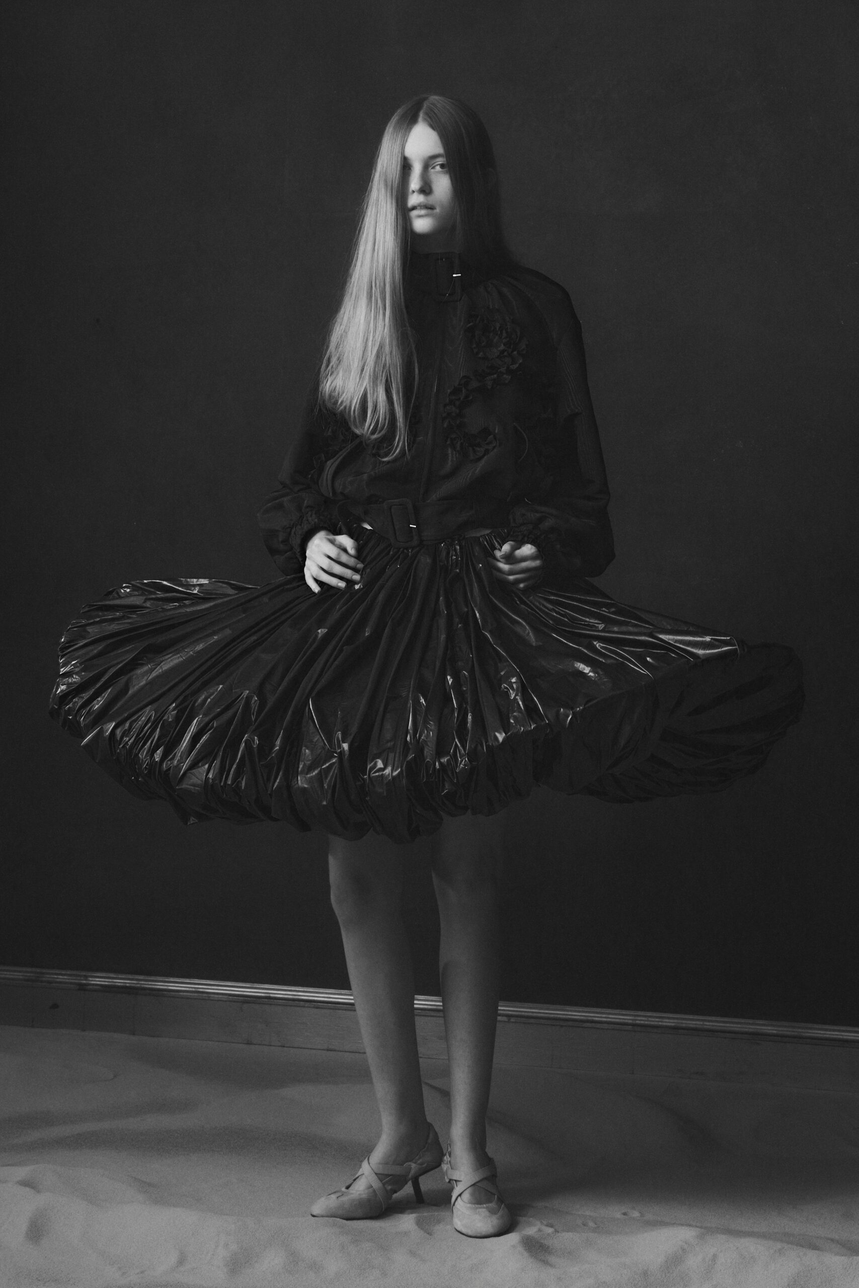 Black high-neck dress with a padded vinyl bubble hem and rosette appliqué. Wide self belt, square buckle sitting slightly off center. Pale suede pumps with crossed straps and narrow kitten heels. Standing in a dark parlor while the storm presses at the windows.