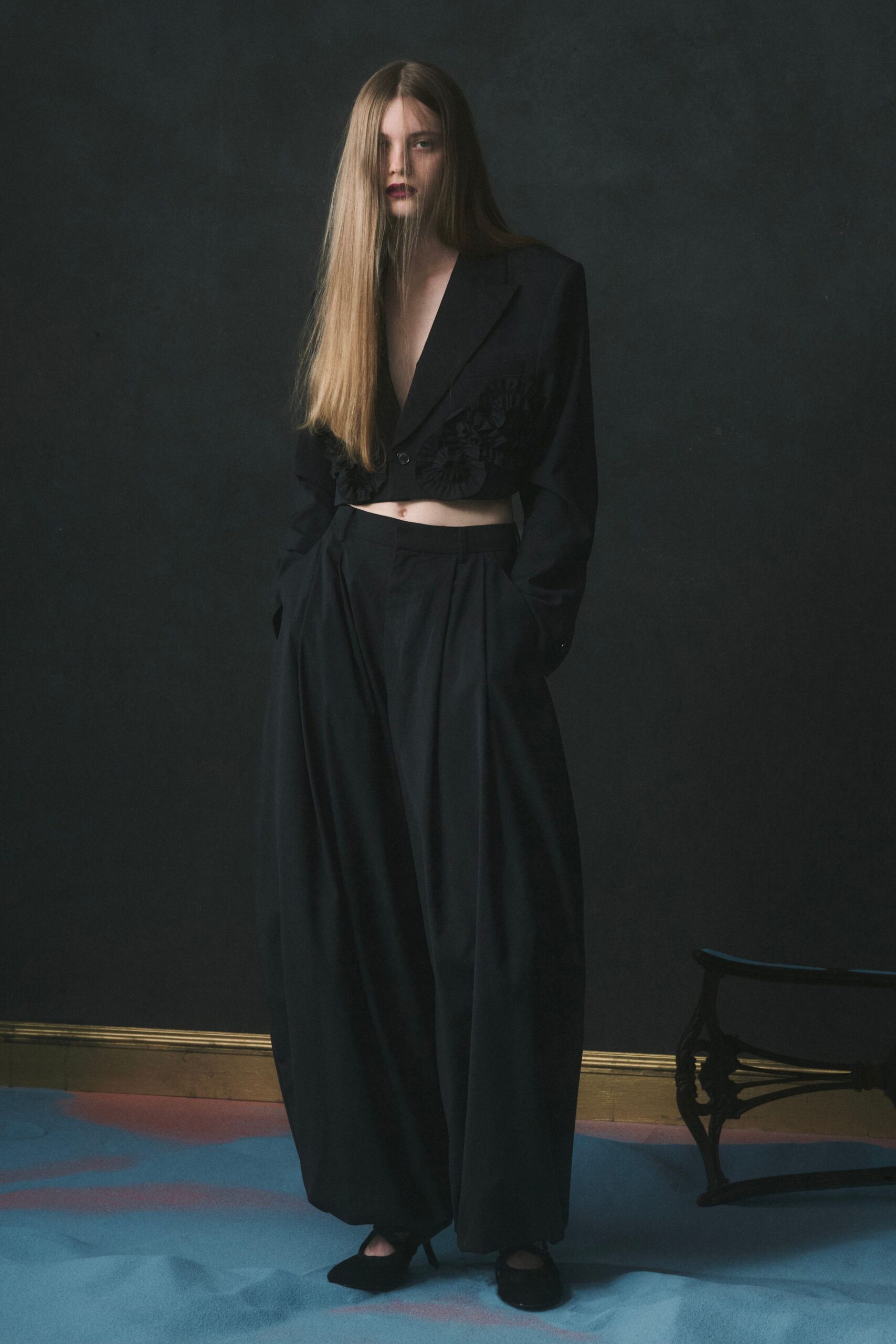 Black cropped blazer with a plunging lapel and rosette appliqué at the hem. Voluminous black trousers, double-pleated and pooling into a rounded break. Black pointed heels with a low curved vamp. After midnight in a townhouse salon, someone has opened the piano bench.