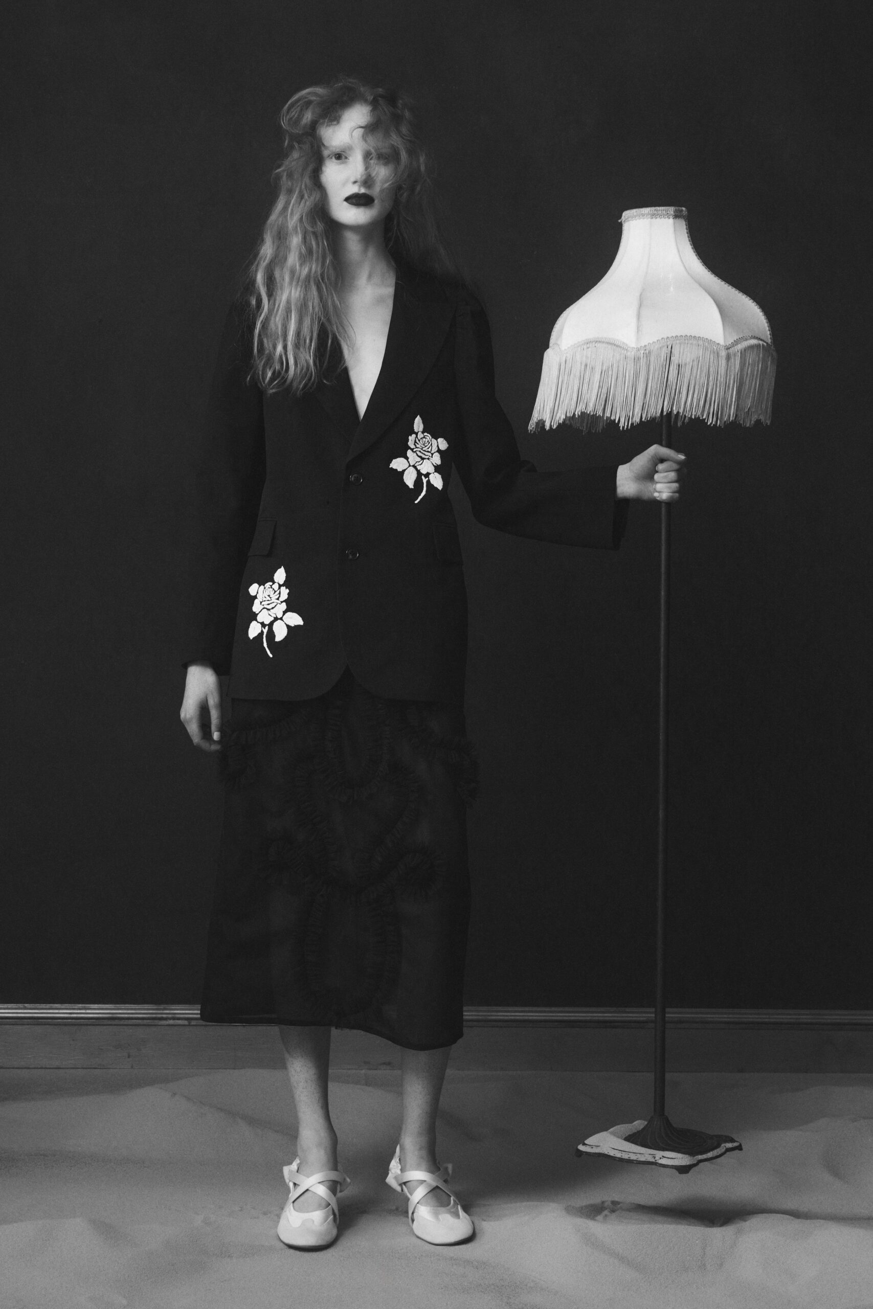 Black tailored blazer with white rose appliques climbing one hip and chest. Calf-length black skirt traced with looping ruffled cord across the surface. White crisscross strap flats with rounded toes. Holding the parlour lamp steady after everyone else has gone upstairs.