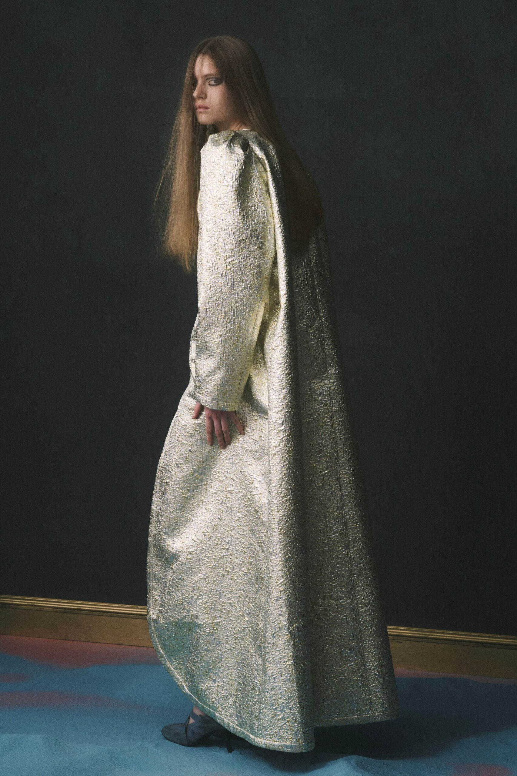 Floor-length silver lamé gown with a cape back and puckered metallic surface. Dark satin heels with crossed straps peeking under the hem. After-hours museum corridor, turning just before the guard says no photos.