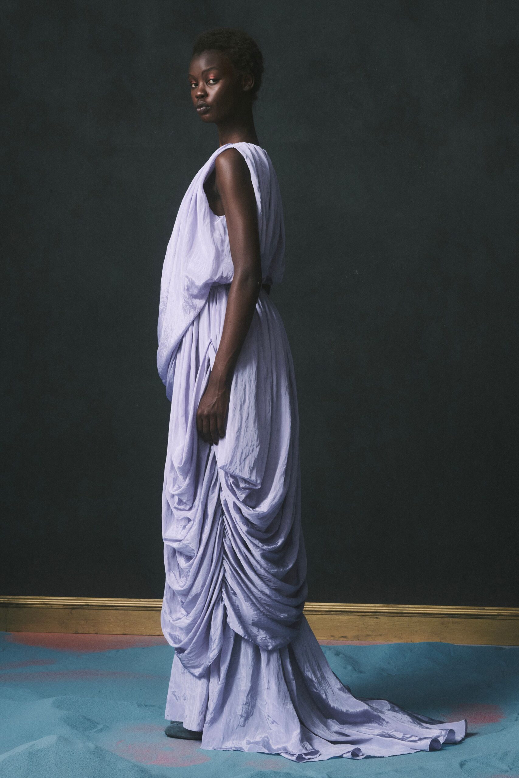 Pale lilac dress in crinkled satin, bodice draped loose over a black belt. Matching skirt heavily ruched into low swags, one central gather pulling longest. Museum hush after closing, shoes soft against the painted floor.