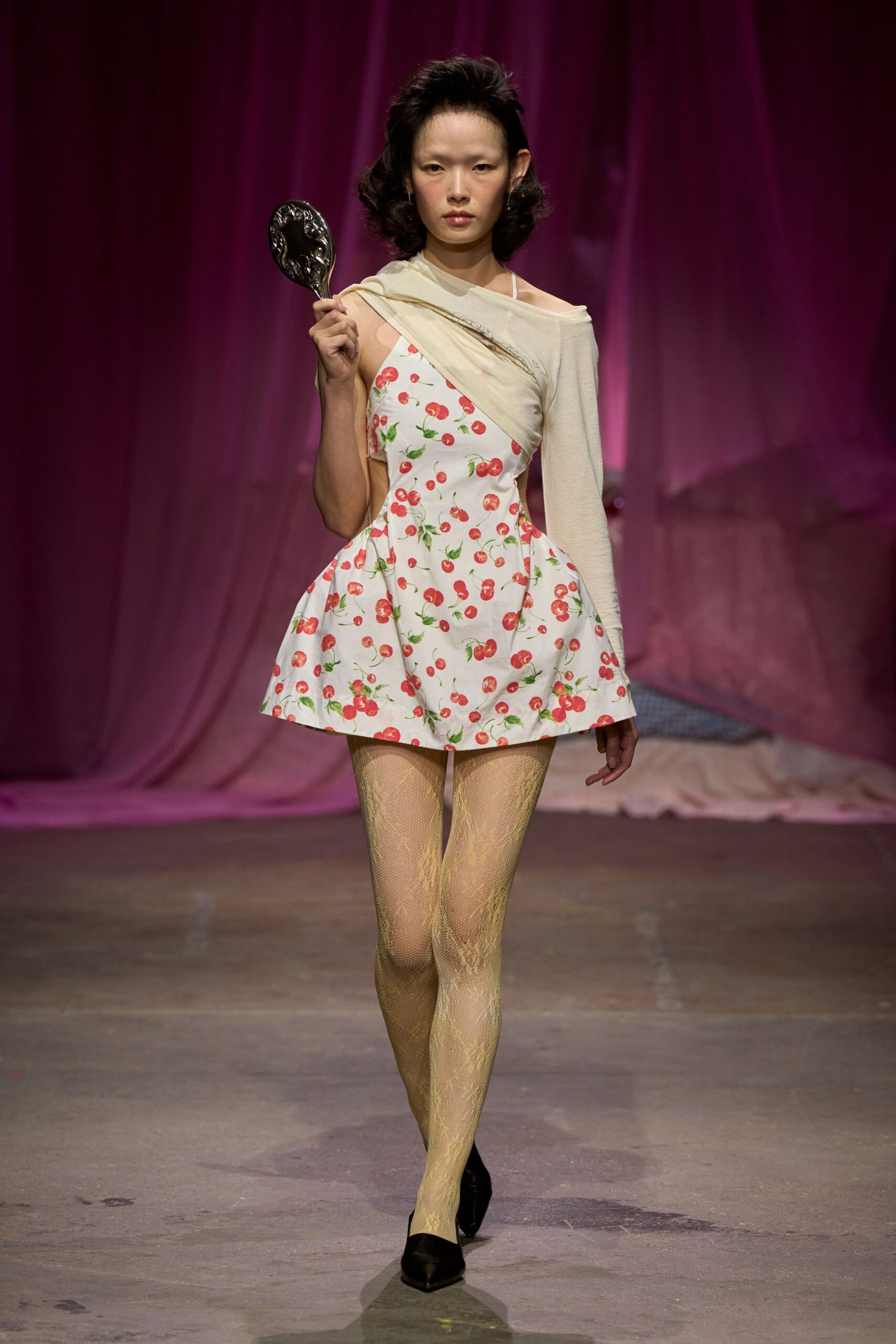 Ornate black hand mirror carried upright by the handle. White fit-and-flare minidress scattered with glossy red cherries. Cream jersey panel twisted off one shoulder, one long sleeve. Pale gold snake-lace tights. Black satin pointed pumps. Checking your face in a fairground mirror after the sun has gone down.