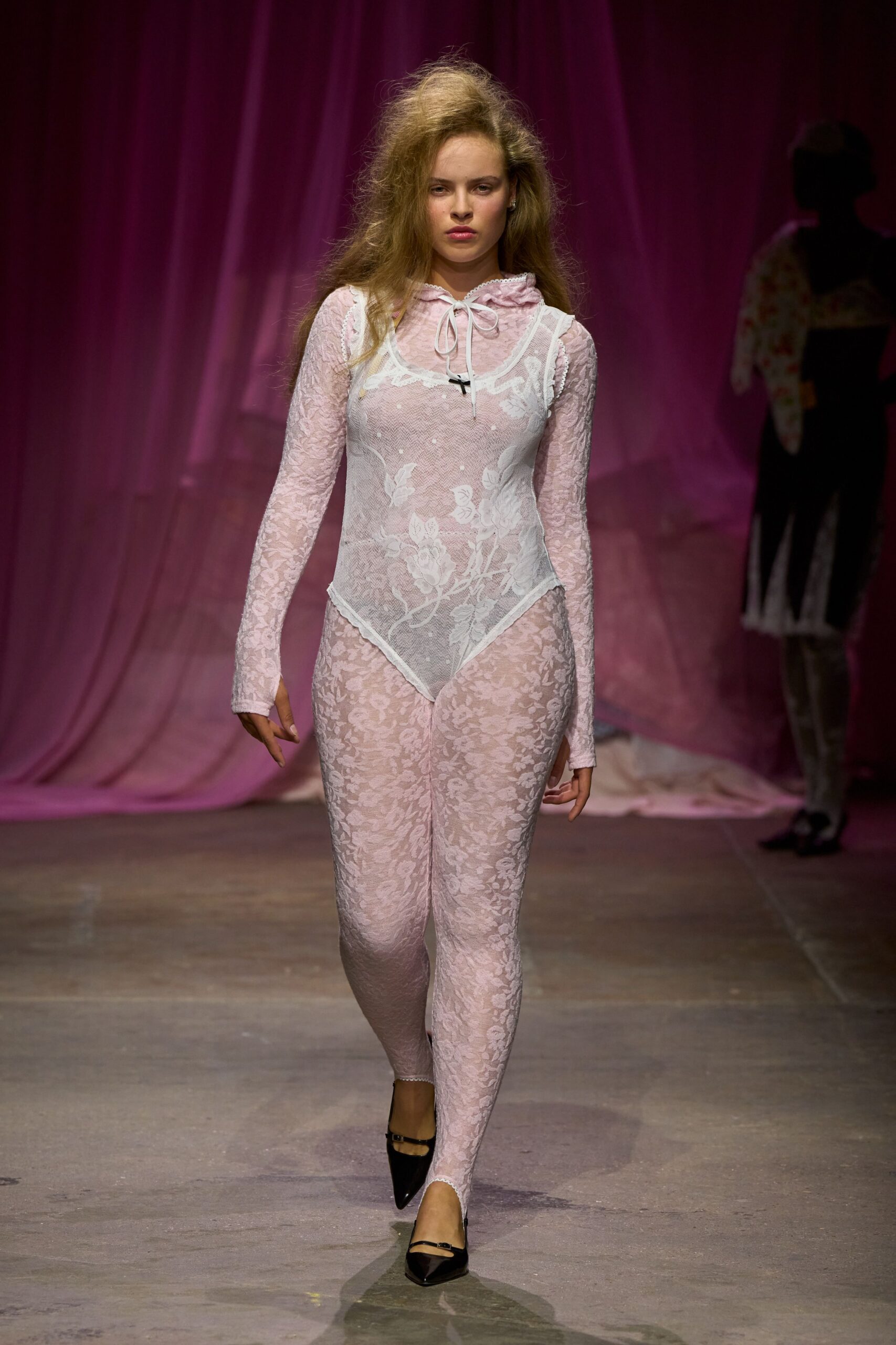 Powder-pink floral lace catsuit, hood tied at the throat with white ribbon. Sheer white mesh bodysuit layered over it, appliqued with larger lace blooms. Black patent Mary Janes with a thin strap across the instep. Walking home from a costume party after sunrise, makeup still intact.