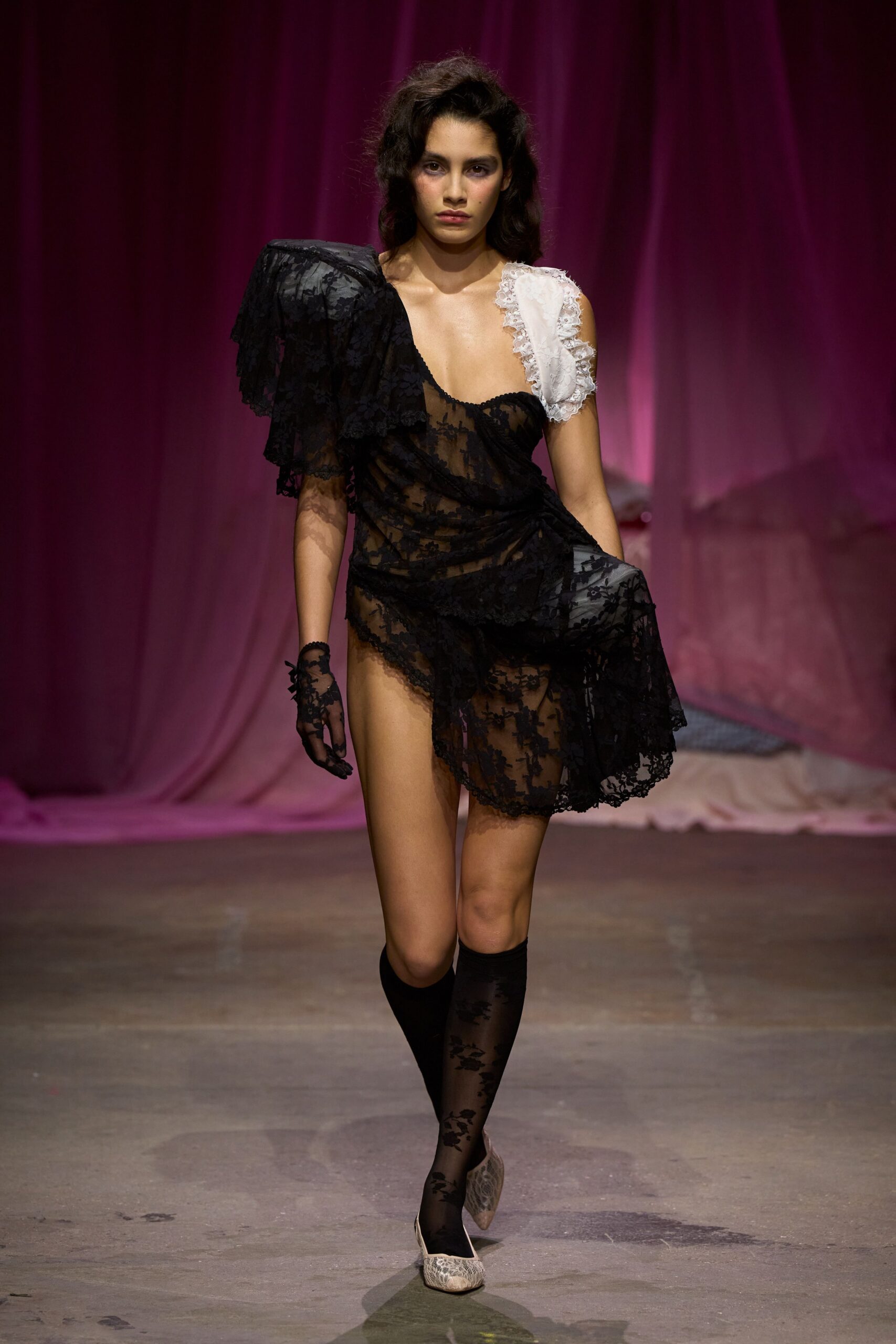 Sheer black floral lace dress, one shoulder stacked with drooping ruffles. White lace-edged strap padded like a little pillow. Single black lace glove, fingers ending just past the knuckles. Black floral mesh knee socks. Pale snakeskin pumps with pointed toes. Whatever you pull on after the motel wedding ran long and strange.