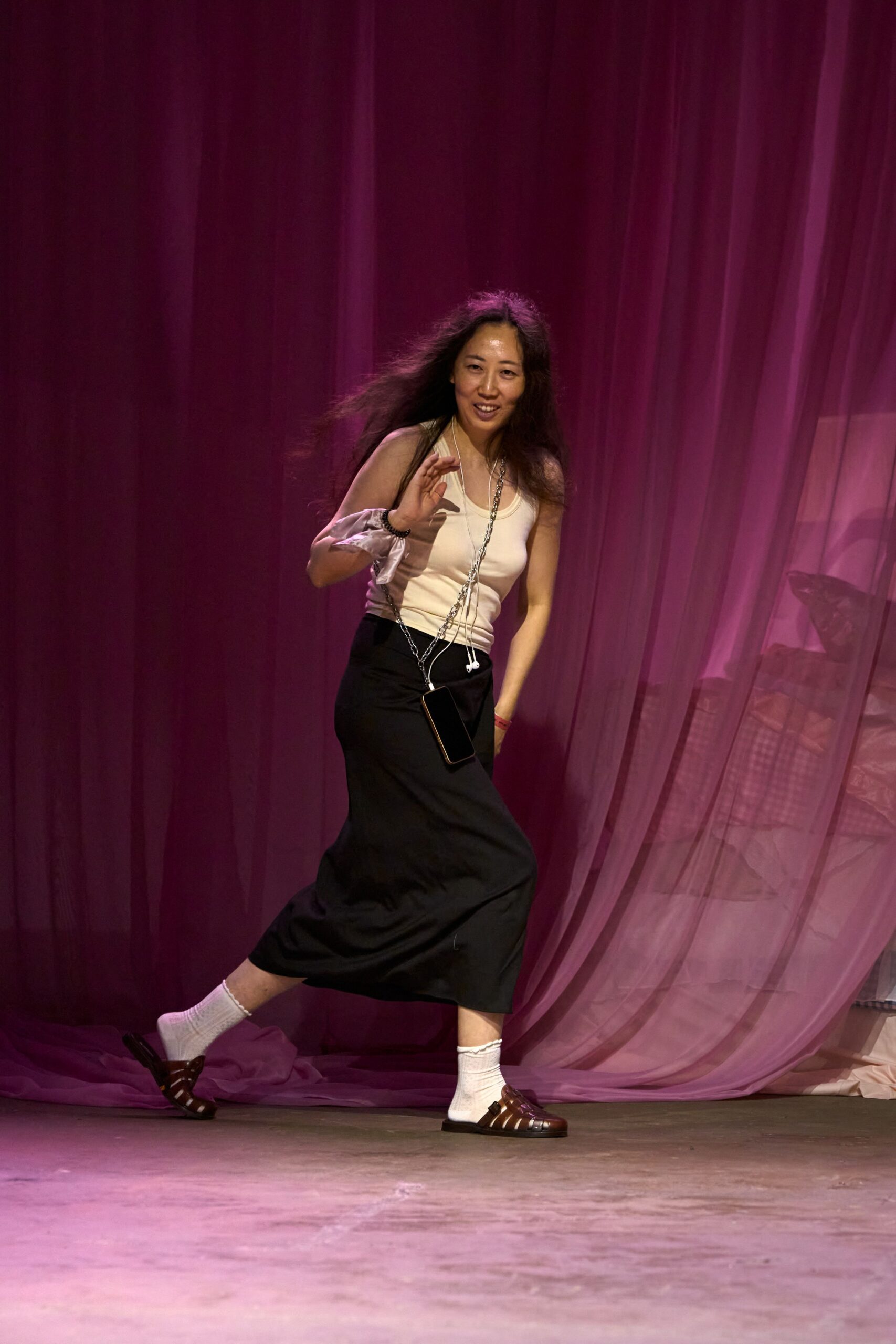Cream ribbed tank, cut close with a scooped neckline. Black phone hanging on a long silver chain crossbody. Black bias-cut midi skirt, hem kicking out at mid-step. White ribbed crew socks with a lettuce-edge cuff. Brown fisherman sandals in glossy leather. Walking across a studio floor while the curtain still sways behind you.