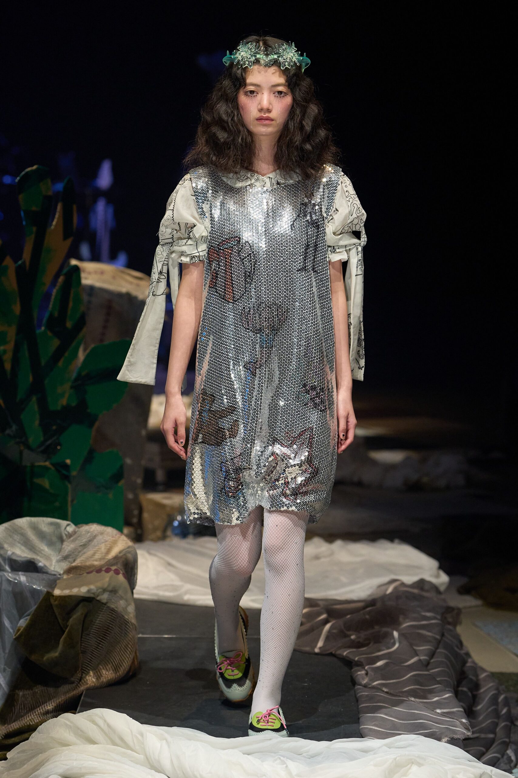 Silver paillette shift dress over a white printed blouse with ruched sleeves tied into long streamers. White fishnet tights, saddle shoes with pink laces, translucent floral headband. Like a school play in a damp church hall.