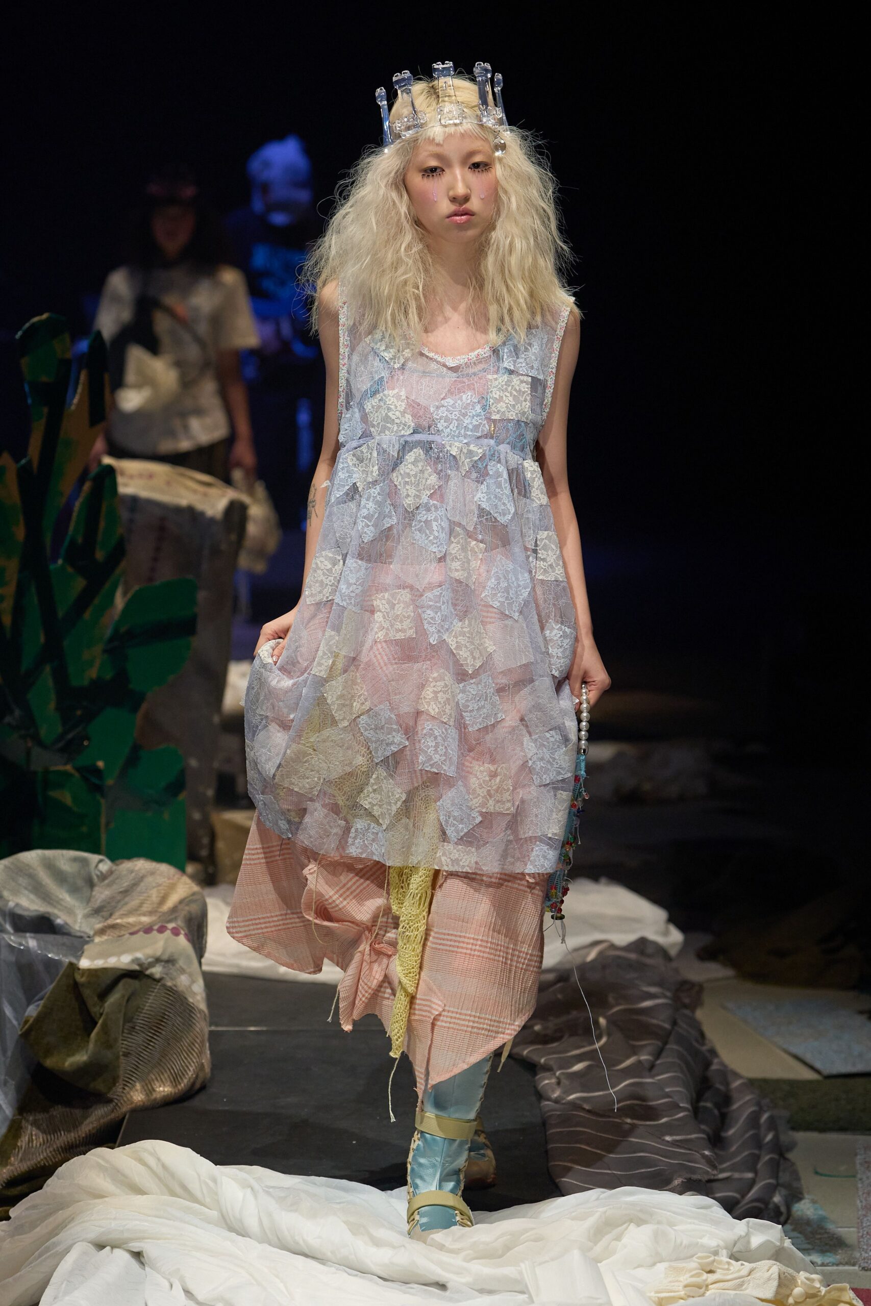 Sheer powder-blue mesh pinafore dress covered in appliqued lace squares, worn over a blush check underlayer with a loose knot at the shin. Pale blue satin boots strapped in beige. Backstage at a school play gone ghostly.
