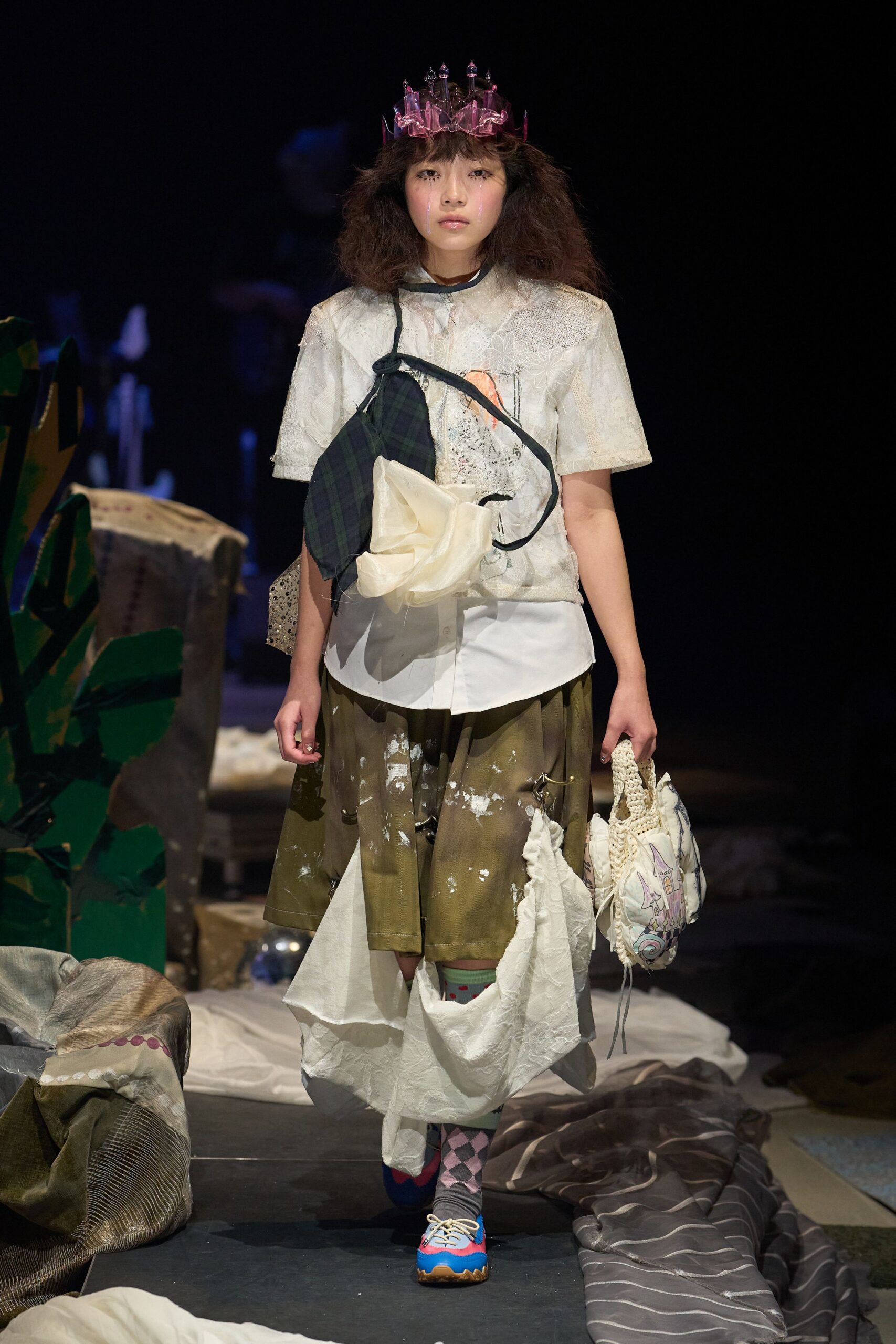Ivory lace-patched short-sleeve top over a white shirttail hem, crossed by a tartan strap with a crumpled organza bloom. Olive skirt splashed with white paint, drooping cloth panel, mismatched socks, blue lace-ups. Like leaving craft camp after dark.