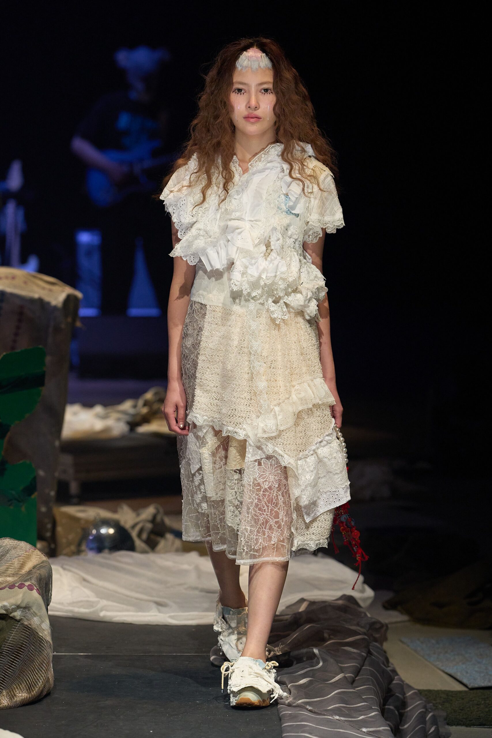 Ruffled ivory blouse in broderie anglaise and lace, the neckline collapsing softly off one shoulder. Patchwork cream lace skirt with a sheer floral net hem. Scuffed trainers with loose laces. Like drifting through a half-stripped hotel room at dawn.