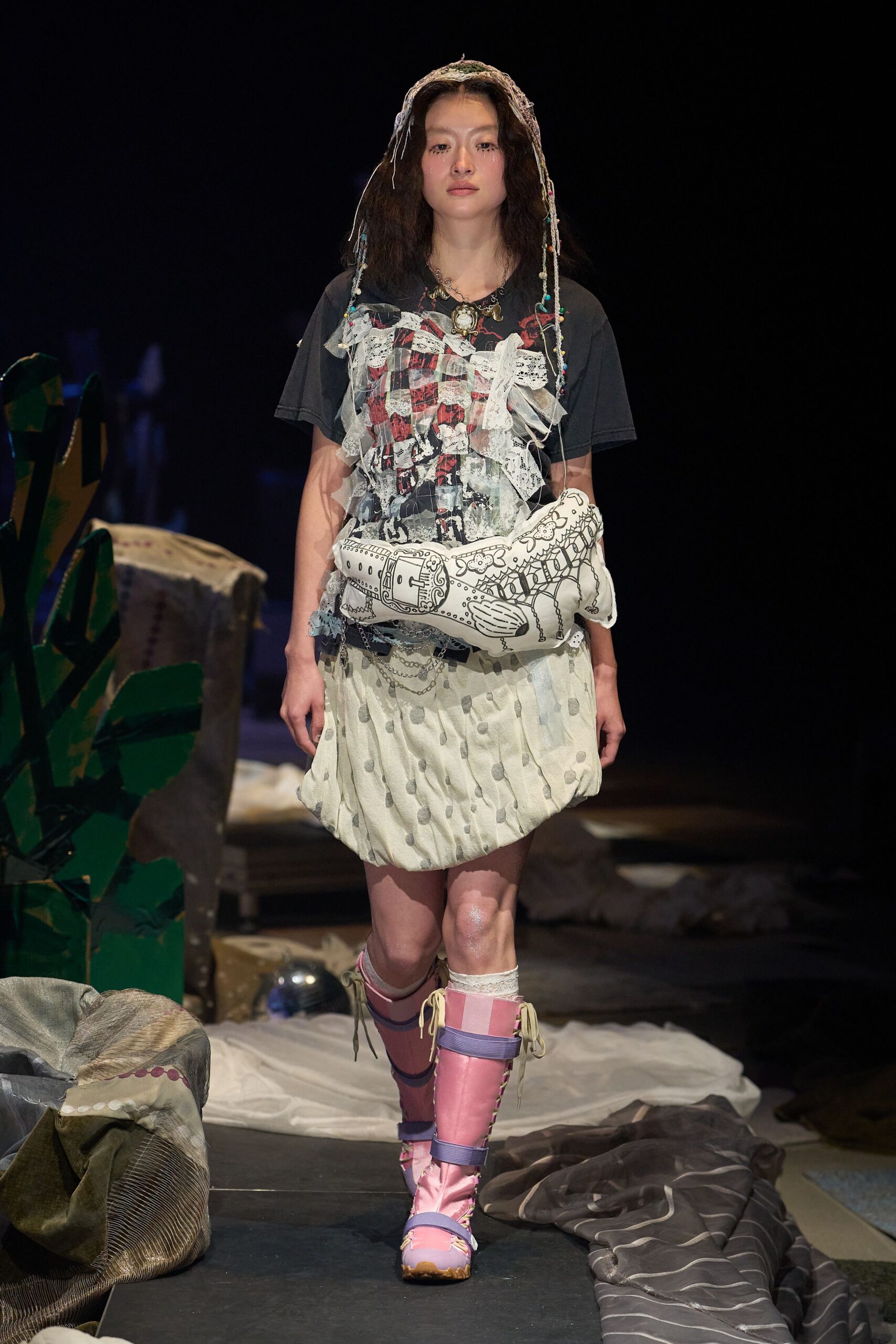 Black graphic T-shirt veiled in shredded tulle and lace scraps, cream bubble skirt with clipped polka dots, illustrated pillow bag tucked at the hip, pink satin lace-up boots. Backstage at a school play gone feral.