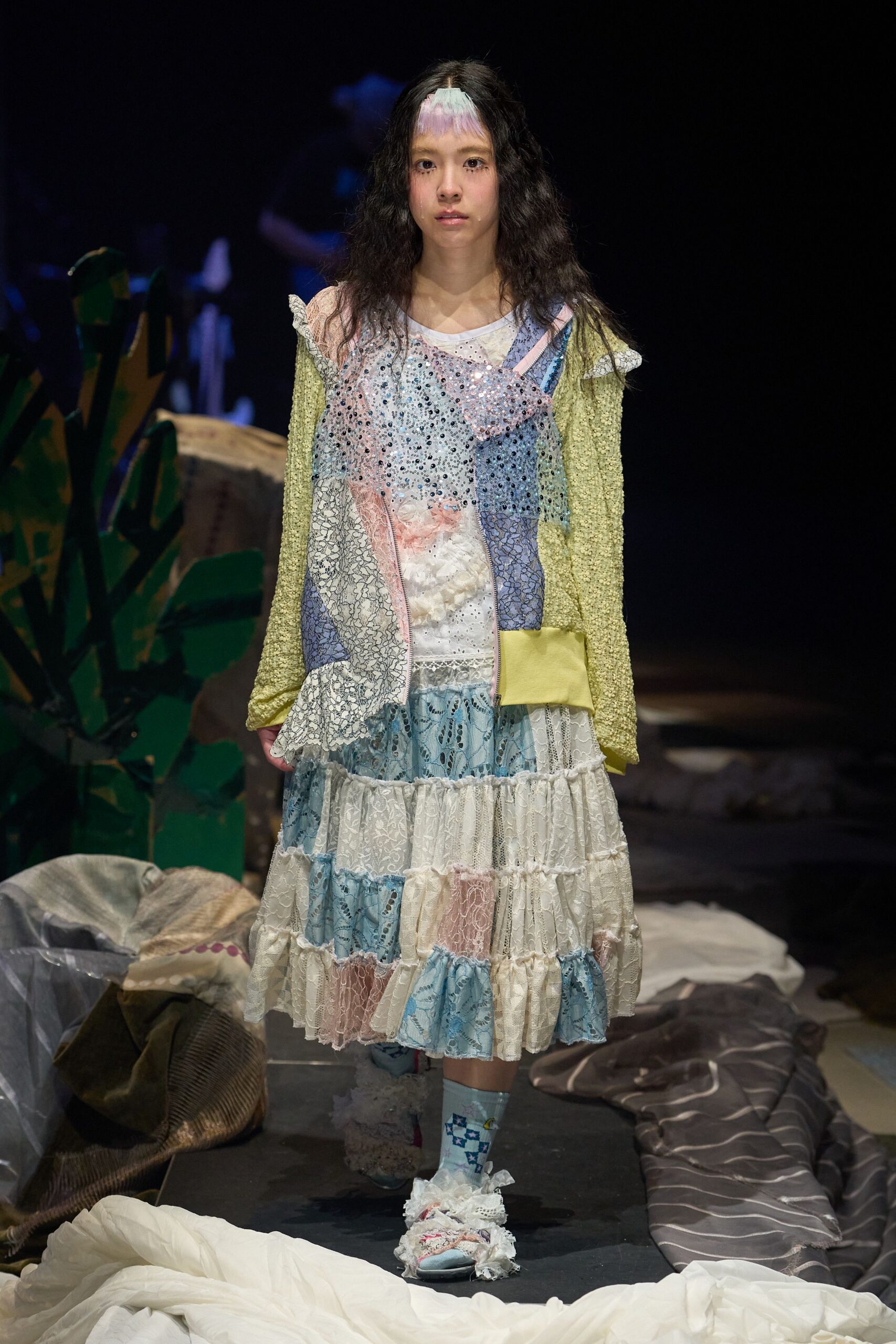 Pale bouclé cardigan with satin cuffs over a pastel patchwork slip, sequins scattered across the bodice. Tiered lace dress beneath, blue intarsia socks, frilled slippers. Like dawn in a holiday rental after everyone else is asleep.