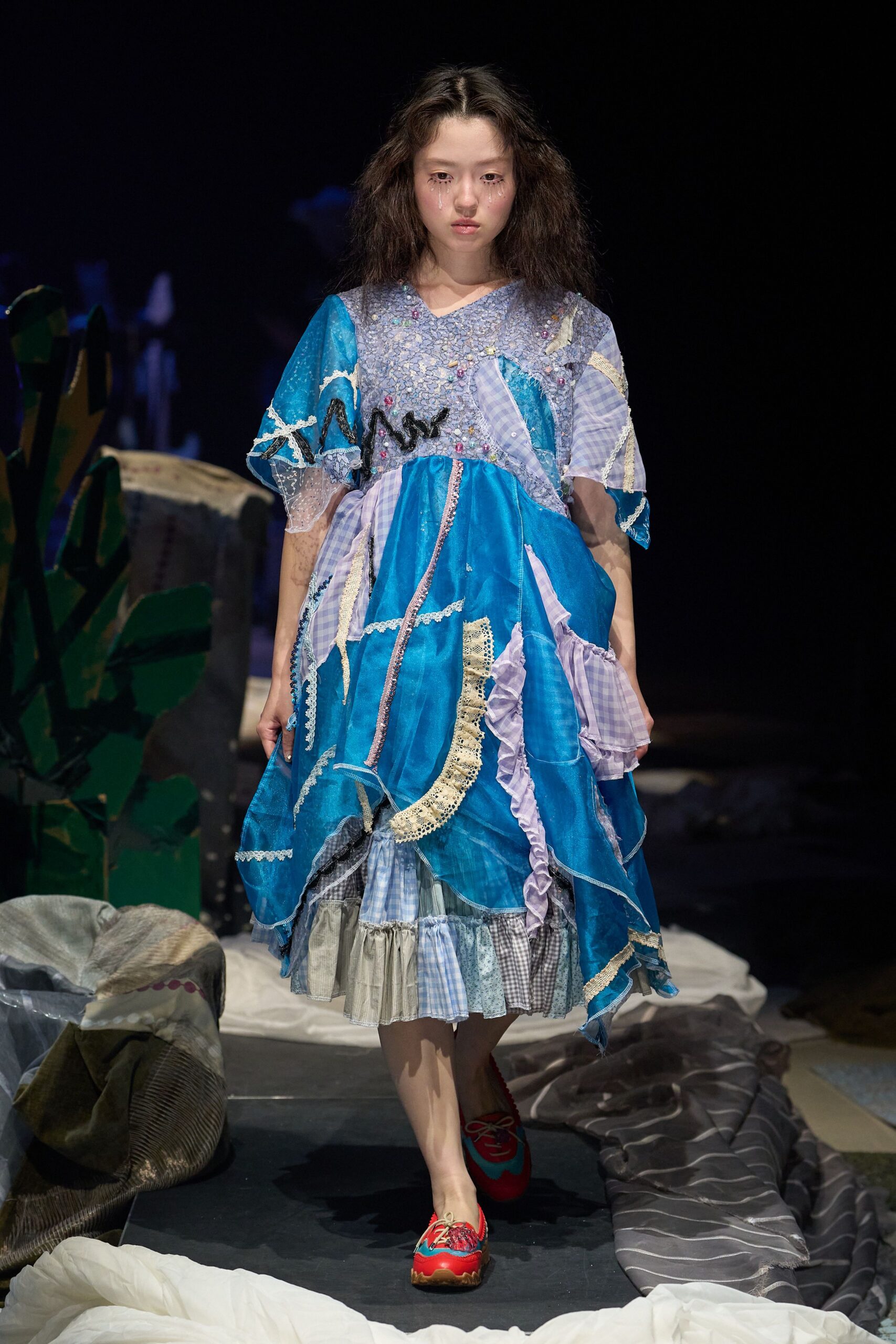 Patchwork dress in cobalt organza and lilac gingham, pieced with crochet lace and a raw zigzag appliqué across the chest; ruffled hem in mixed checks. Red lace-up flats with chunky tread soles. Like wandering an aquarium after the school trip ends.
