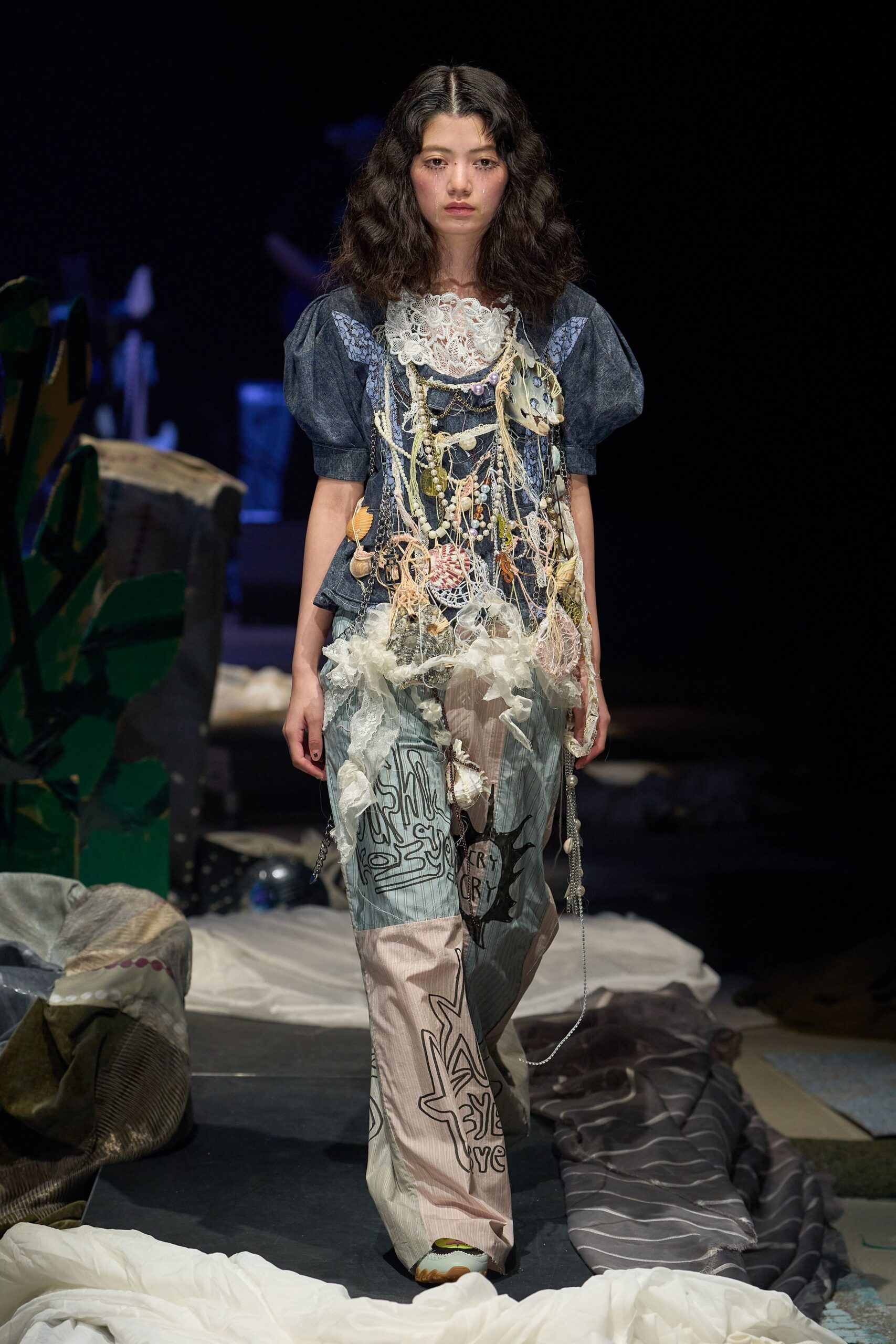 Dark chambray dress with puff sleeves and a dense guipure lace bib, buried under a tangled bib of shells, beads, chains and ripped net. Pale striped trousers with doodled panels. Like beach salvage spread across a rehearsal room floor.