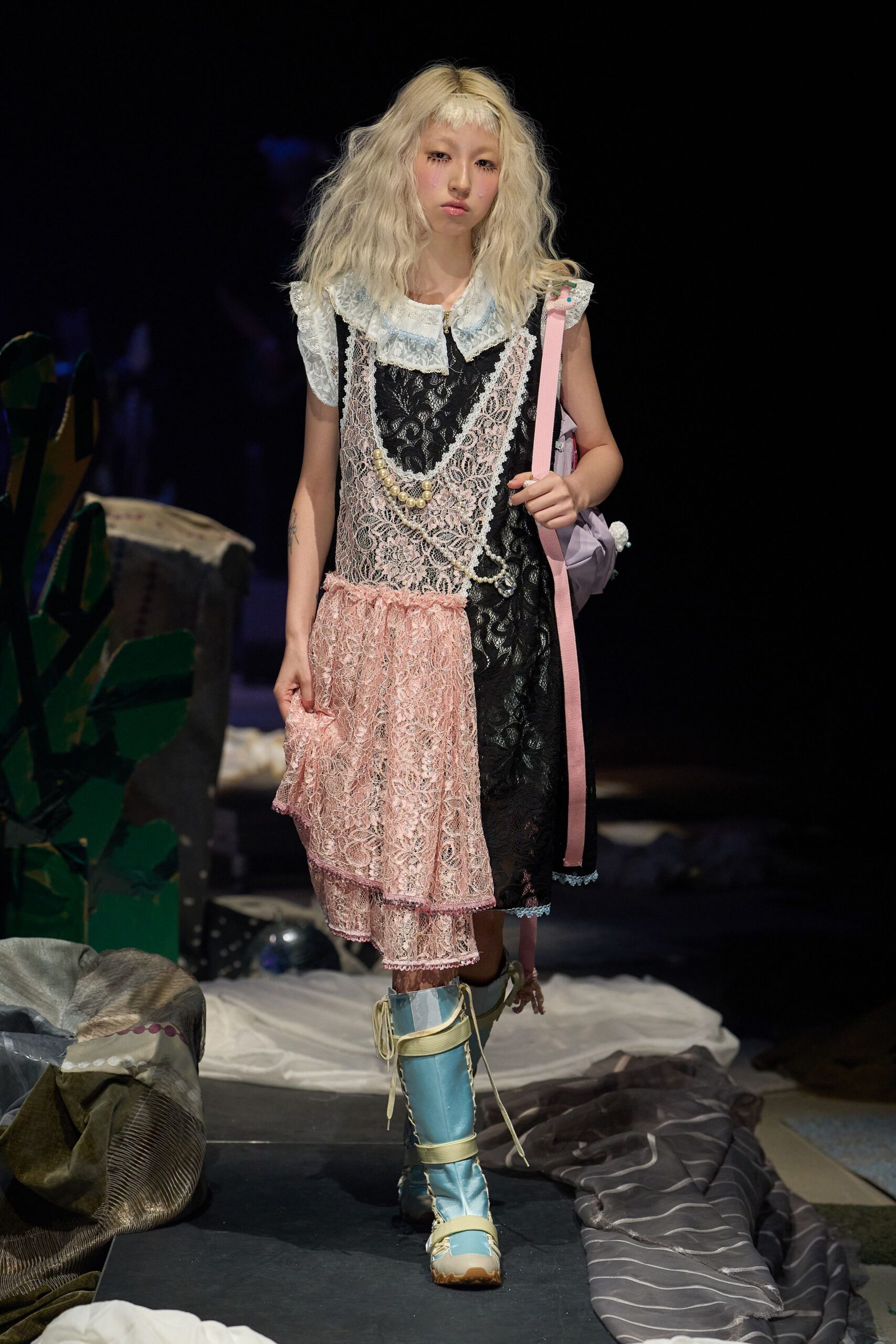 White blouse with a scalloped lace collar under a black floral-lace slip dress, draped in pale pink lace ruffles and a loop of faux pearls. Lilac bag on a grosgrain strap, powder-blue lace-up boots. Like dawn in a cluttered doll studio.