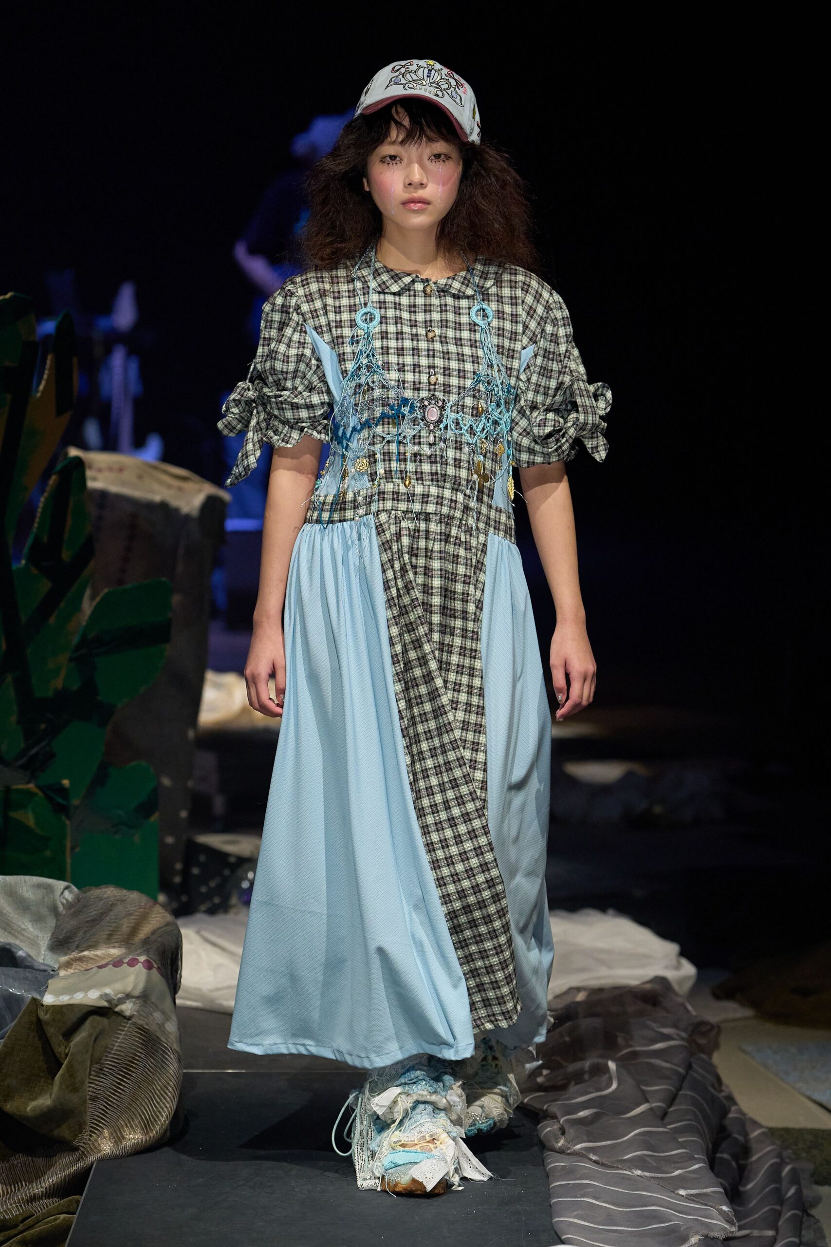 Embroidered white baseball cap, checked dress with ruched tie sleeves and a skewed placket, overlaid with pale blue chiffon panels and a beaded net bib. Lace-wrapped shoes. Like getting dressed from the lost-and-found trunk at a summer camp recital.