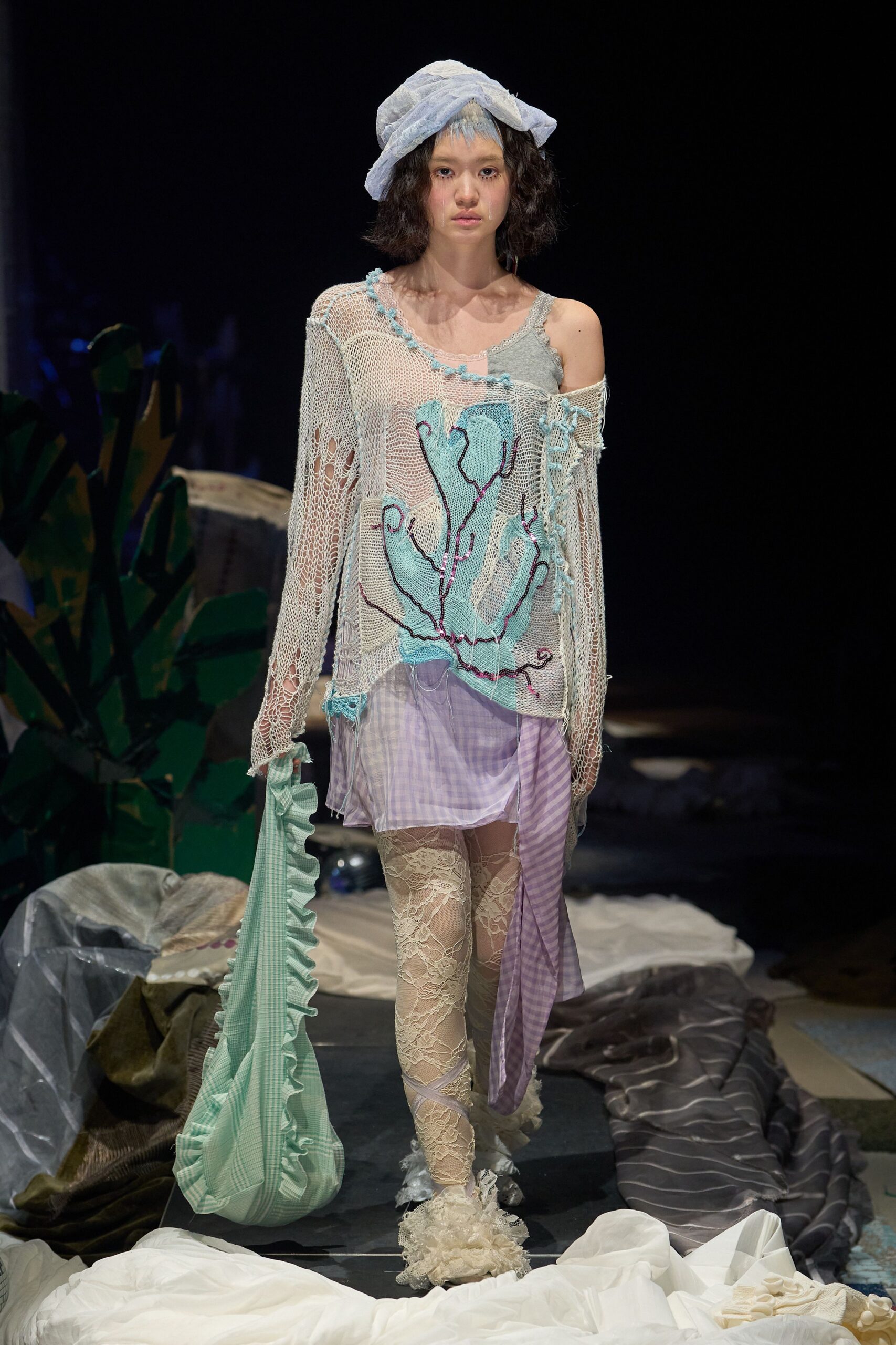 Cracked-open openwork knit slouching off one shoulder, coral-like intarsia picked out in loose purple chain stitch over a grey camisole. Lilac gingham slip skirt, cream lace leggings, crumpled bonnet. Dawn cleanup after a beach wedding.