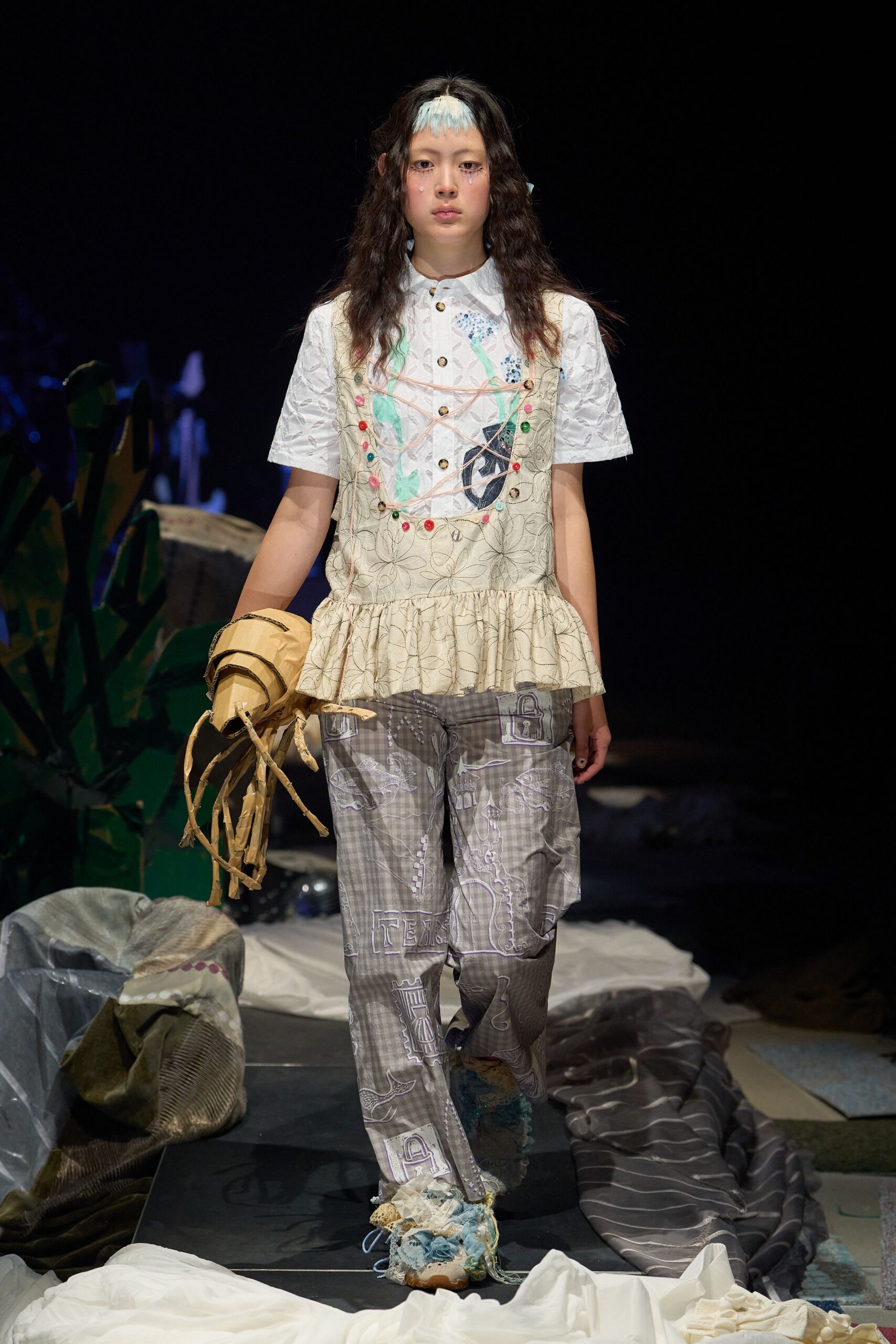 White short-sleeve shirt in puckered jacquard, topped with a cream peplum vest traced in loose floral stitching and pink cord lacing. Grey plaid trousers printed with padlocks, crumpled paper bag in hand. Backstage at a school play gone strange.
