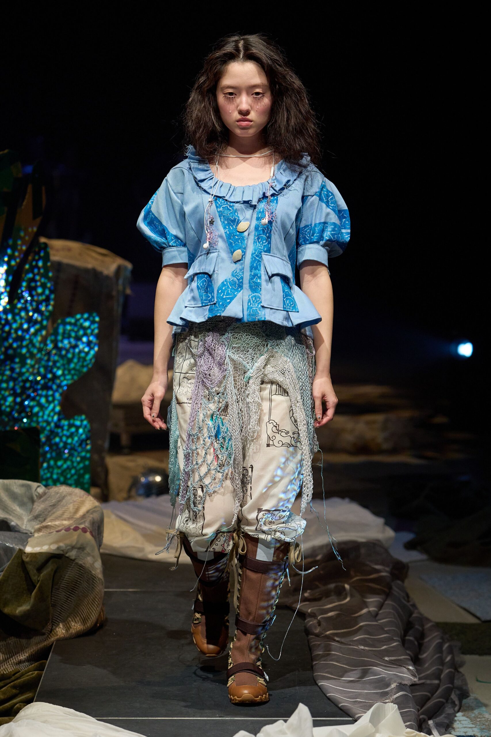 Sky-blue jacket with puff sleeves, a ruffled neckline and flap pockets jutting at the waist. Cream printed trousers draped in loose fishnet. Brown lace-up boots with ribbed shin panels. Backstage at an aquarium after closing.