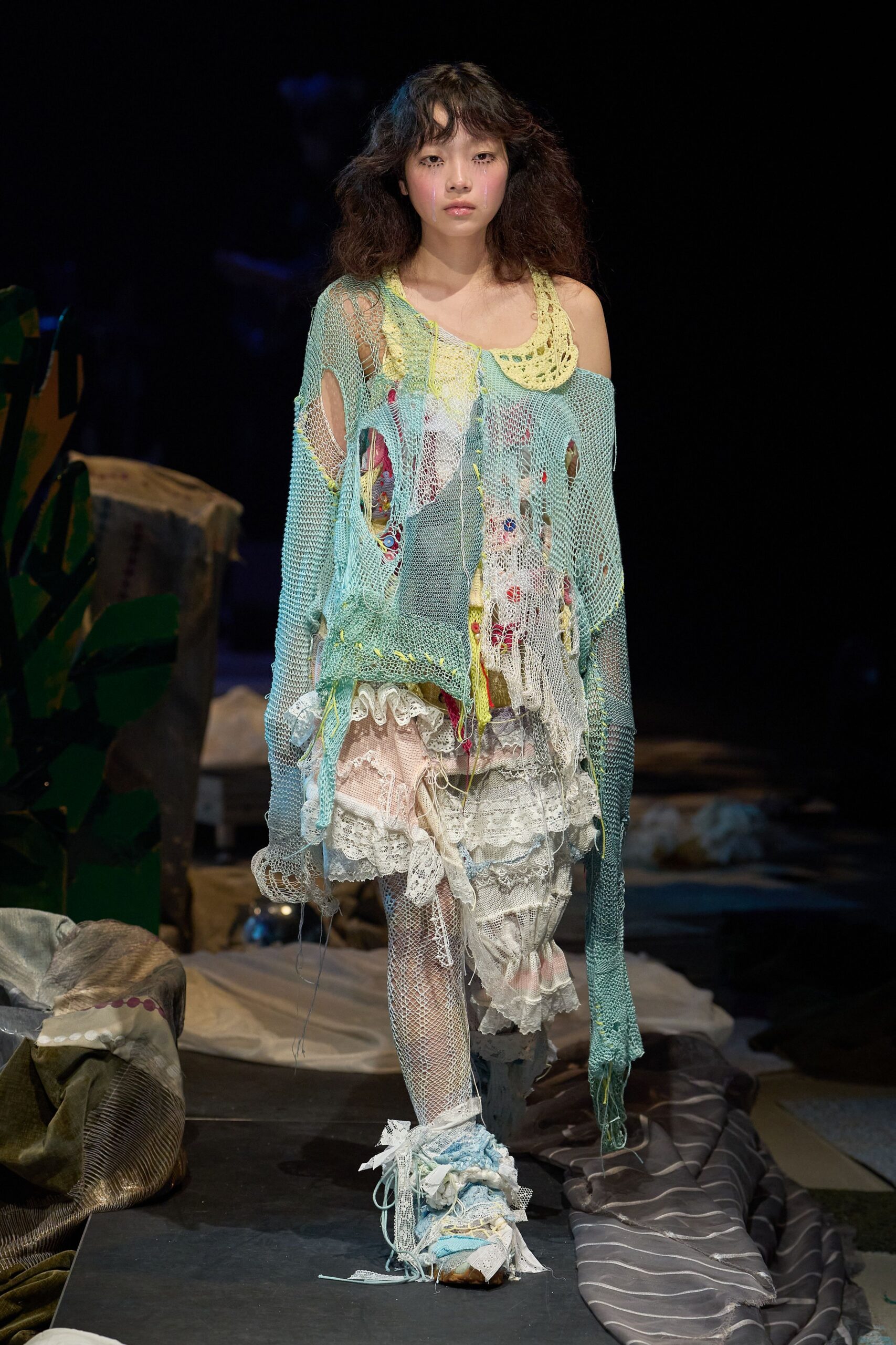 Mint open-knit sweater shredded into long ladders, slipping off one shoulder over a lemon crochet bib. Patchwork slip of lace flounces and mesh stockings, ribbon-wrapped shoes. Like dawn in a cluttered costume studio.