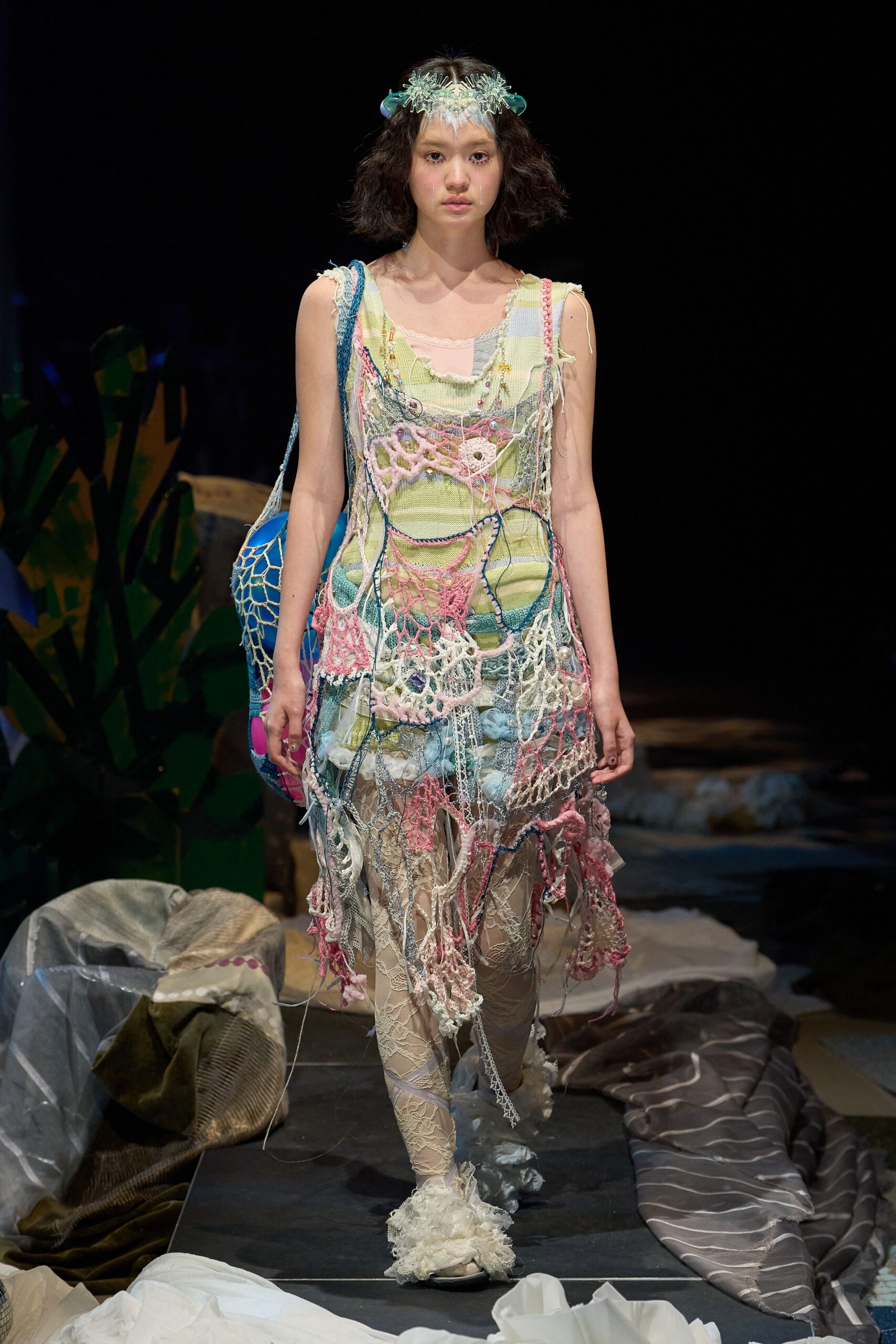 Patchwork slip dress in pale pistachio knit, draped with a shredded crochet net threaded in pink and blue cords. Ivory lace leggings, ruffled slippers, netted orb bag. Like beach salvage brought home in a storm.