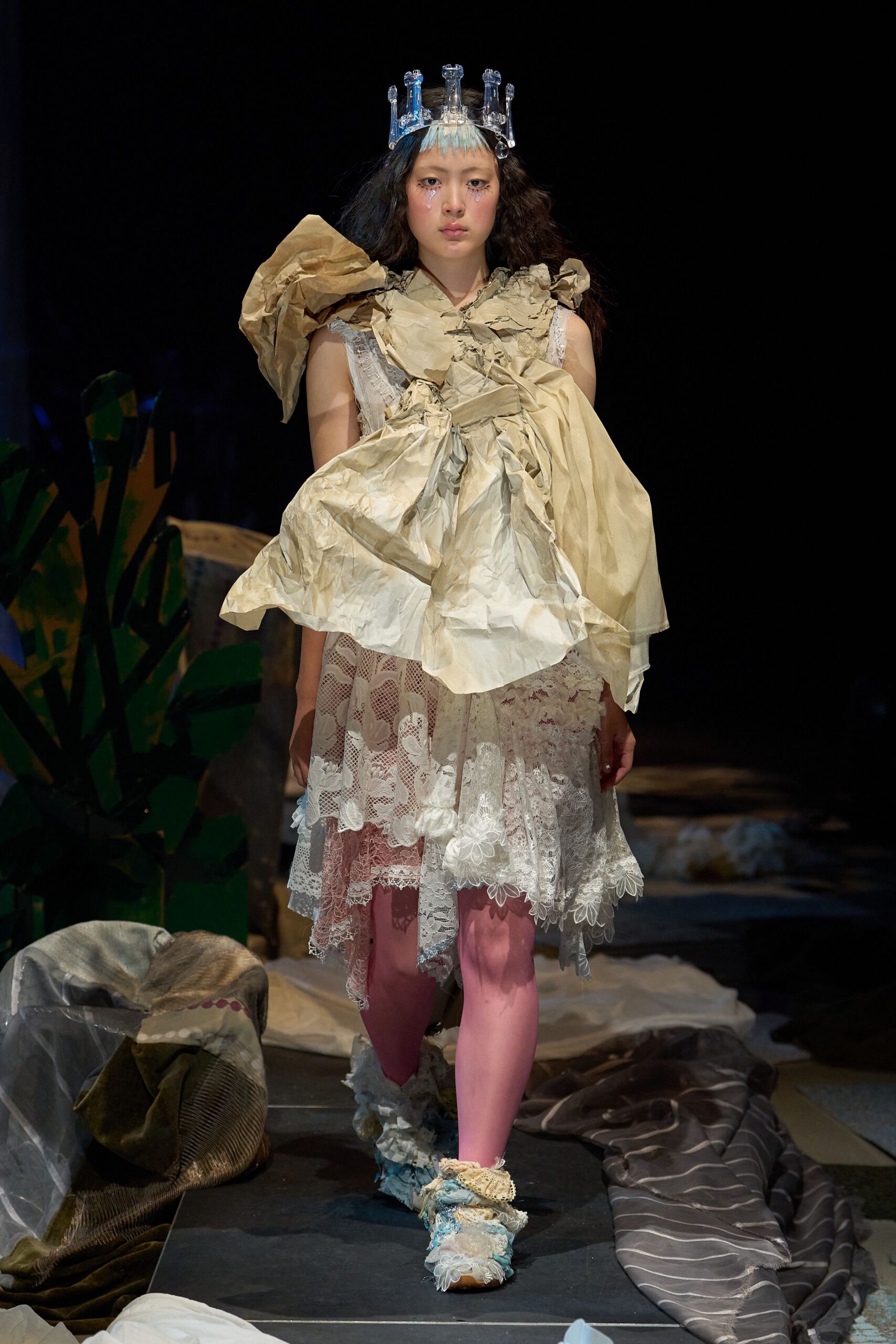 Crumpled parchment-like shoulder wrap knotted into oversized bows over a sheer lace dress with scalloped panels and an uneven handkerchief hem. Pink tights, frilled ankle wraps, clear crown. After the school play, alone in the prop room.