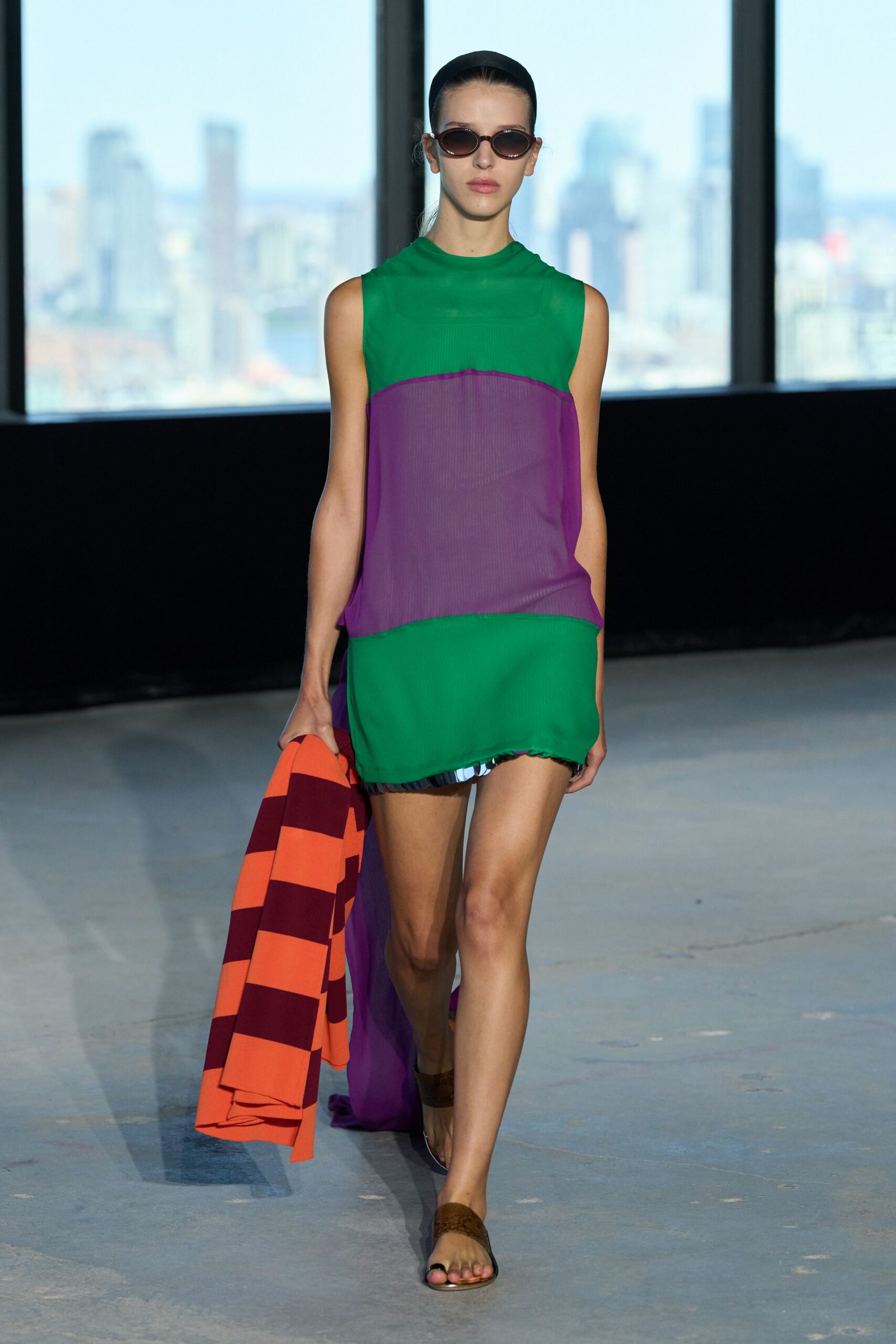 Narrow oval sunglasses with reddish smoked lenses. Sleeveless ribbed dress in emerald and violet sheer panels, a striped underlayer peeking below. Orange coat carried loose, blocked with broad oxblood bands. Flat brown sandals with a wide suede strap. Walking back from a hotel pool after the sun has slipped behind the towers.