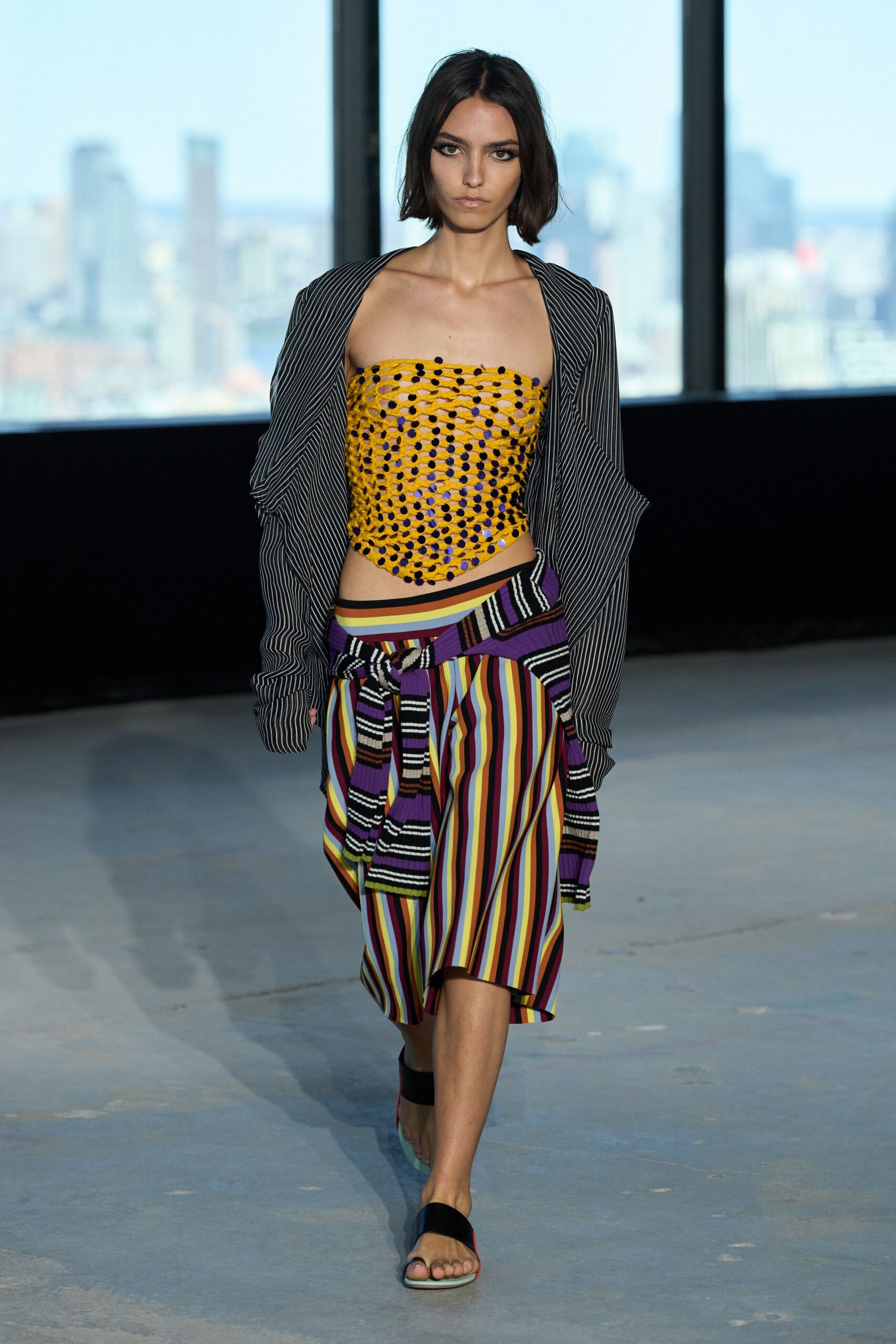 Black pinstripe shrug hanging off the shoulders in soft collapsed folds. Mustard openwork bandeau studded with tiny navy pom-pom knots. Knee-length skirt in dense vertical stripes, purple knit tied at the hips. Black band sandals with a pale mint sole edge. Rooftop laundry hour, city heat still lifting off the concrete.
