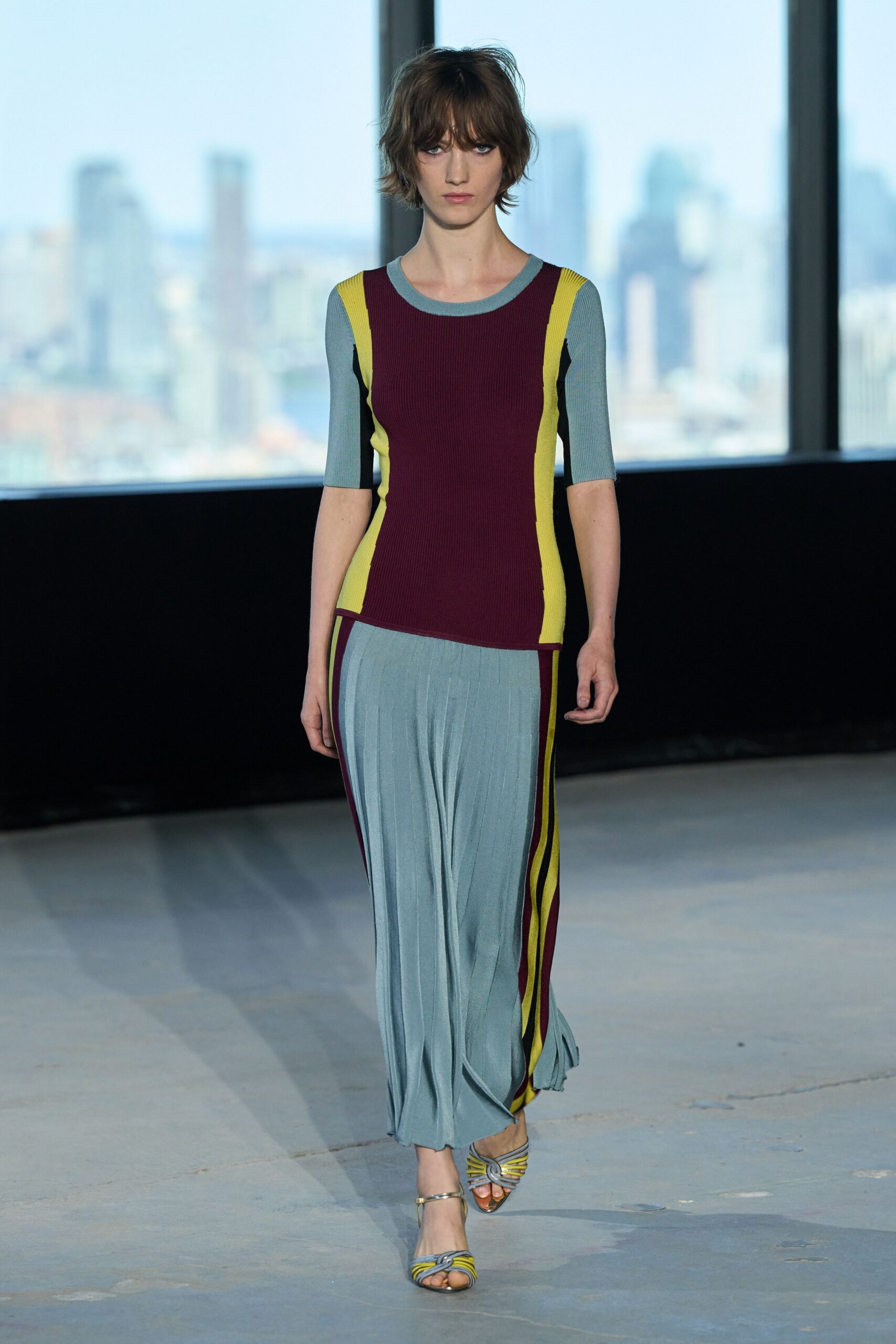 Burgundy ribbed knit top with powder-blue sleeves and chartreuse side panels. Powder-blue ribbed knit skirt falling in long soft vertical folds. Metallic sandals crossed with twisted grey and chartreuse straps. Crossing a glass office tower lobby after sunset, city still lit behind you.