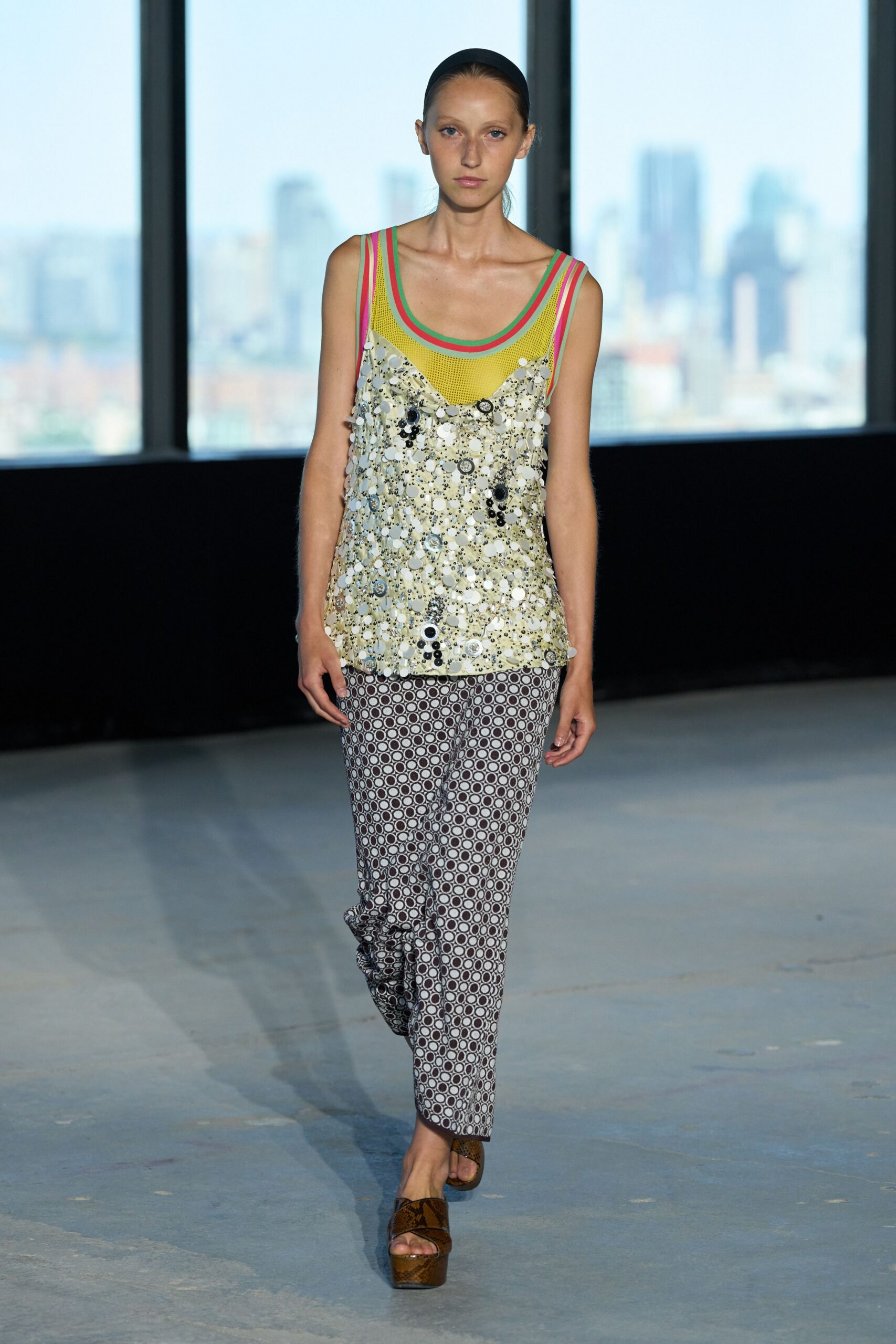 Wide black headband slicking the hair flat. Neon-trimmed yellow mesh tank under a sequinned slip, discs catching light. Slim cropped trousers in tight brown-and-white ring print. Brown python platform slides. Walking to a hotel breakfast after sleeping in last night's makeup.