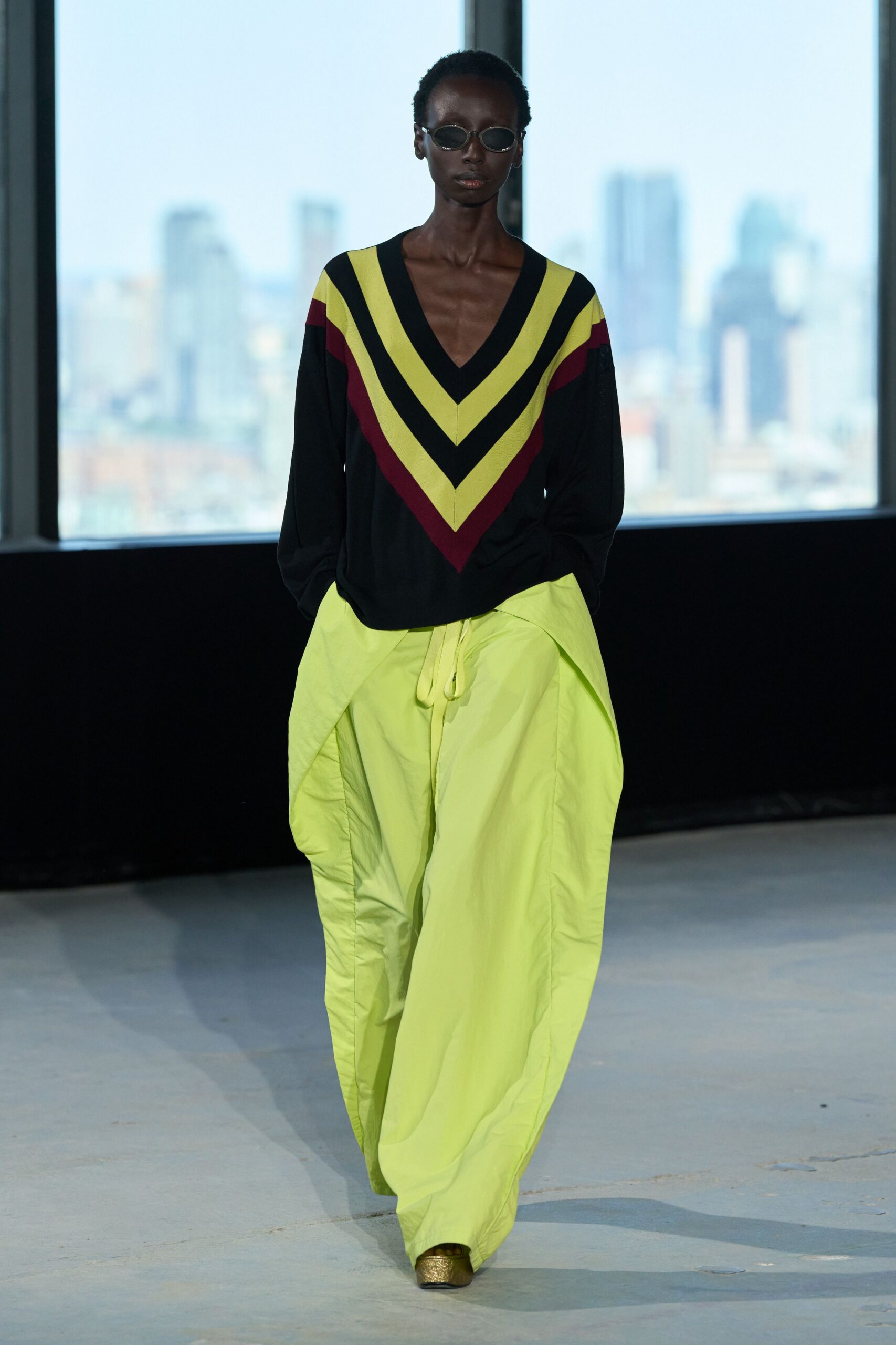Tiny oval sunglasses with dark lenses and a thin metal rim. Black deep-V sweater marked by citron and oxblood chevrons. Acid-lime parachute trousers, gathered by a long dangling drawcord. Gold shoes peeking out under the pooling hem. Crossing a glass office lobby after rain, carrying the whole room in color.