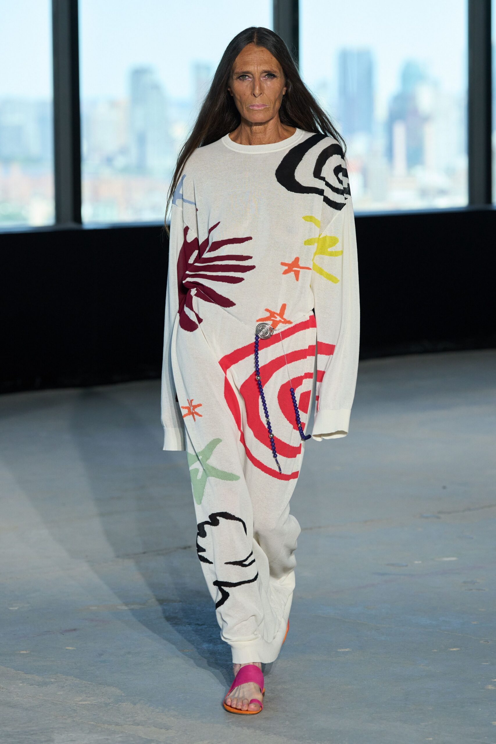 Fine ivory knit with oversized abstract intarsia and slouched extra-long sleeves. Matching knit trousers, gathered at the waist with a silver rosette and blue beaded cords. Fuchsia thong sandals with a sharp orange sole edge. Sunday museum café, children still chalking spirals on the pavement outside.