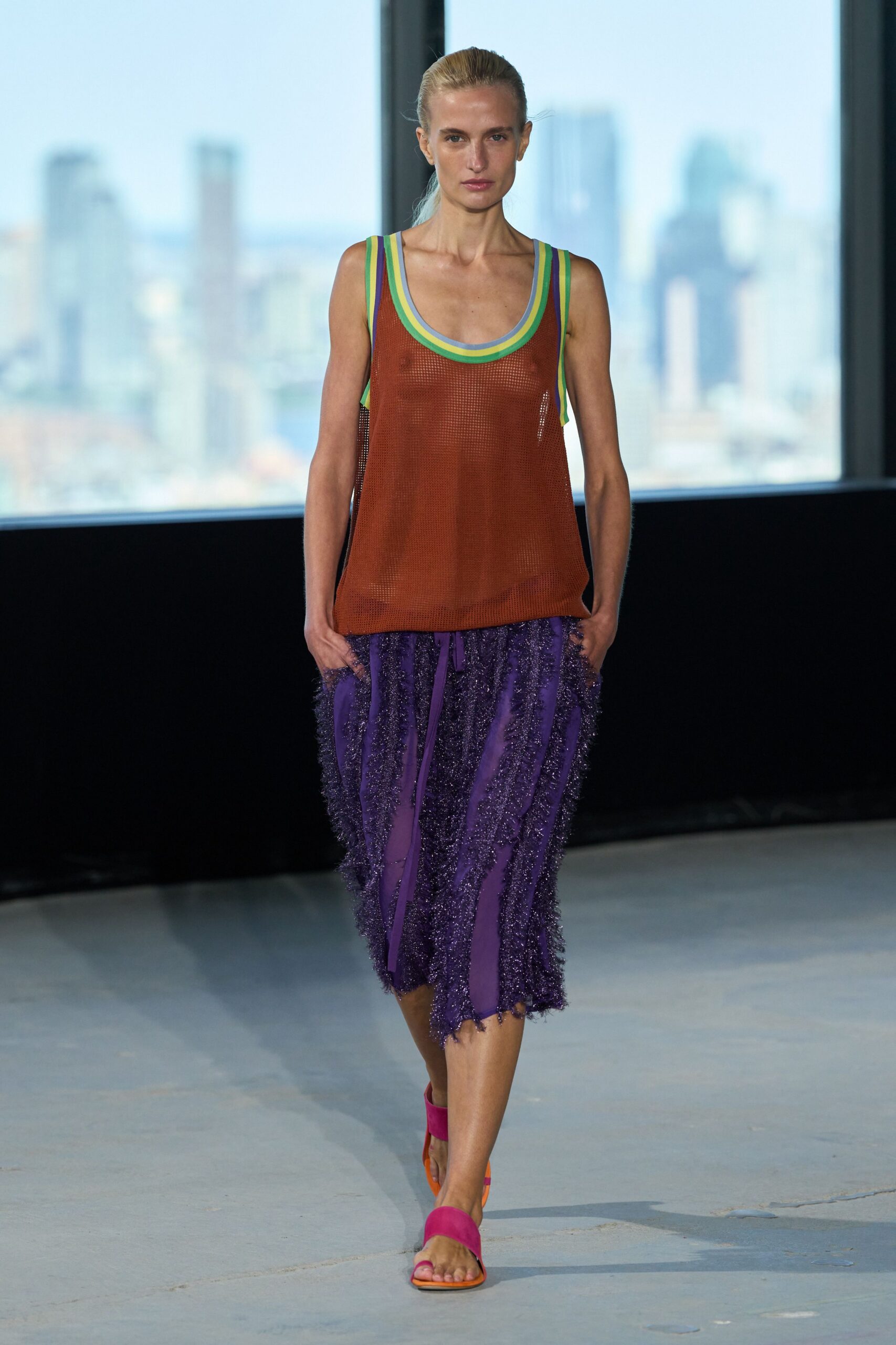 Rust open-mesh tank with layered mint, citron and powder-blue binding. Purple midi skirt paneled with shaggy tinsel fringe, hem hanging uneven. Fuchsia suede bands on bright orange soles. Walking a hotel rooftop at dusk before the pool lights switch on.