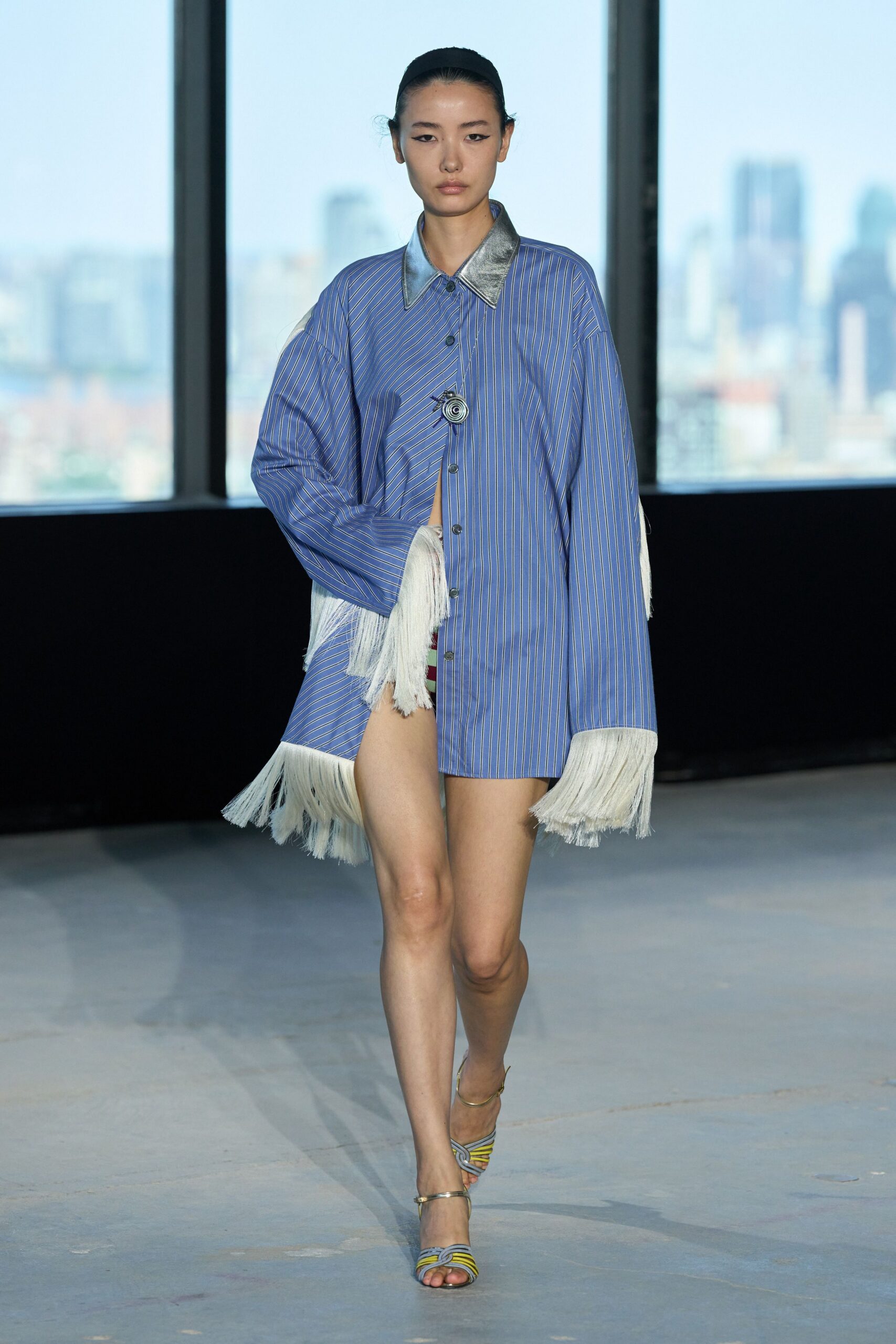 Oversized blue pinstripe shirt dress with a silver pointed collar and long white fringe. Spiral rosette brooch fastening the placket off center. Striped knotted sandals with slim metallic ankle straps. Crossing a hotel atrium just after the pool closed.