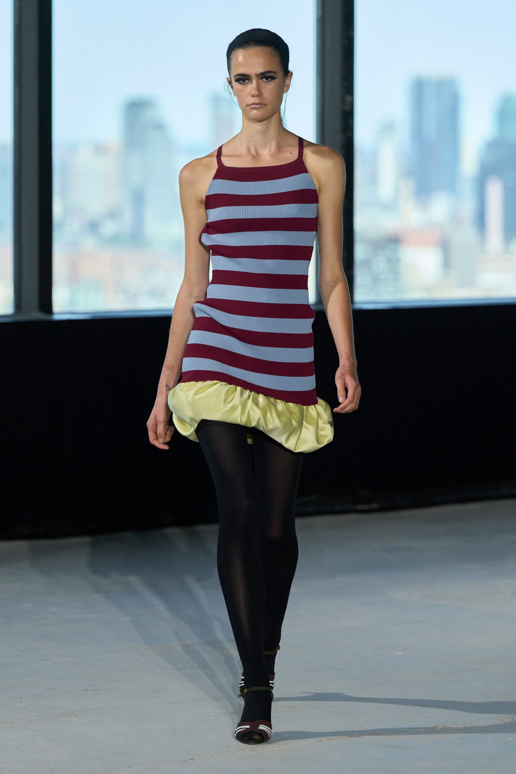 Wide black band holding the hair flat off the face. Burgundy and powder-blue rib knit slip dress with a puffed chartreuse satin hem. Opaque black tights. Burgundy open-toe sandals with white curved straps. Hotel conference room at sunrise, skyline still blurred behind the glass.