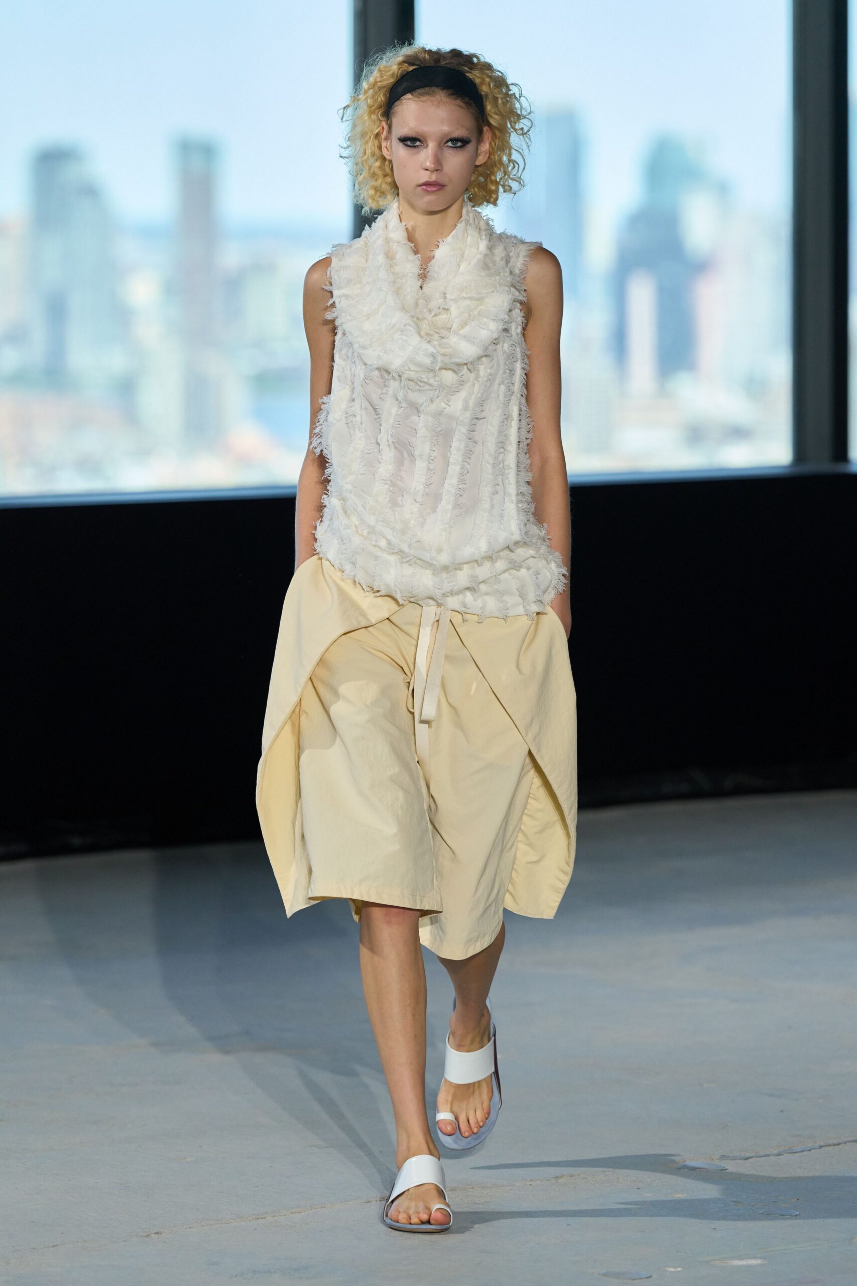 Wide black jersey headband holding back bleached curls. Ivory sleeveless top covered in feathery frayed strips and a soft cowl. Buttercream paper-touch shorts with outsized folded side panels and dangling ties. White leather toe-ring slides. Crossing a skybridge at noon with the river blown pale below.