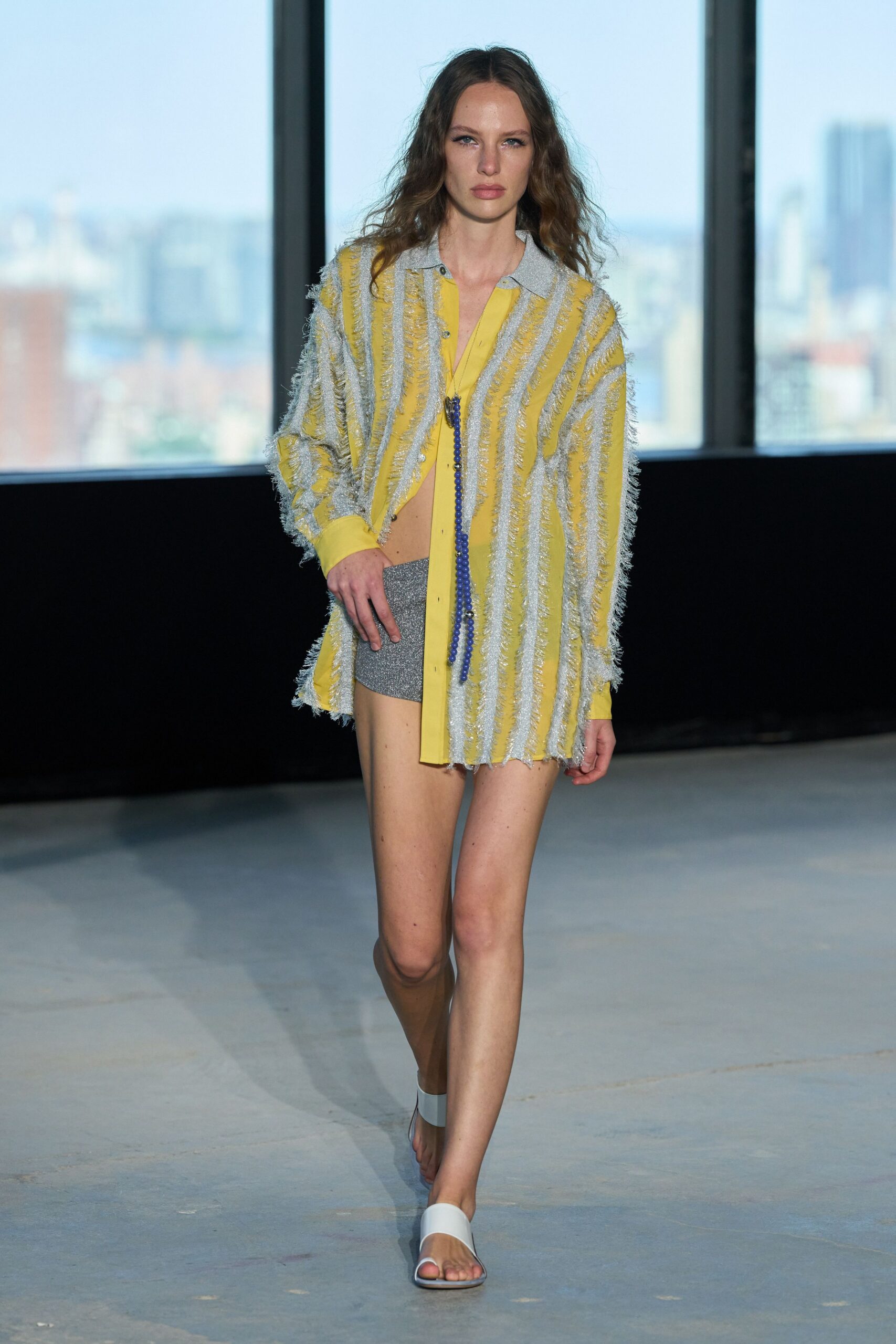 Lemon shirt striped with silver tinsel fringe, placket hanging open past the navel. Long cobalt bead strand dropped straight through the open front. Silver glitter hot pants cut high on the thigh. White slide sandals with a broad curved strap. Roof-deck brunch after rain, wind still lifting the table paper.