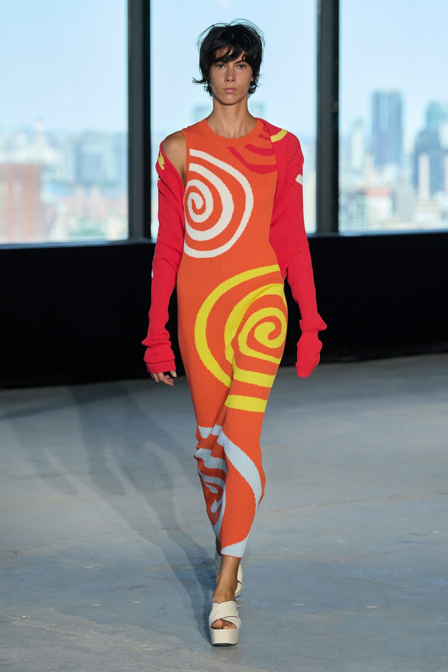 Orange rib-knit jumpsuit with white and citron spiral intarsia curling over one leg. Hot-pink ribbed shrug slipping off one shoulder, sleeves pooling past the hands. White leather platform mules with a broad twisted front strap. Heat shimmer on a rooftop court, somebody chalking lines before sunset.