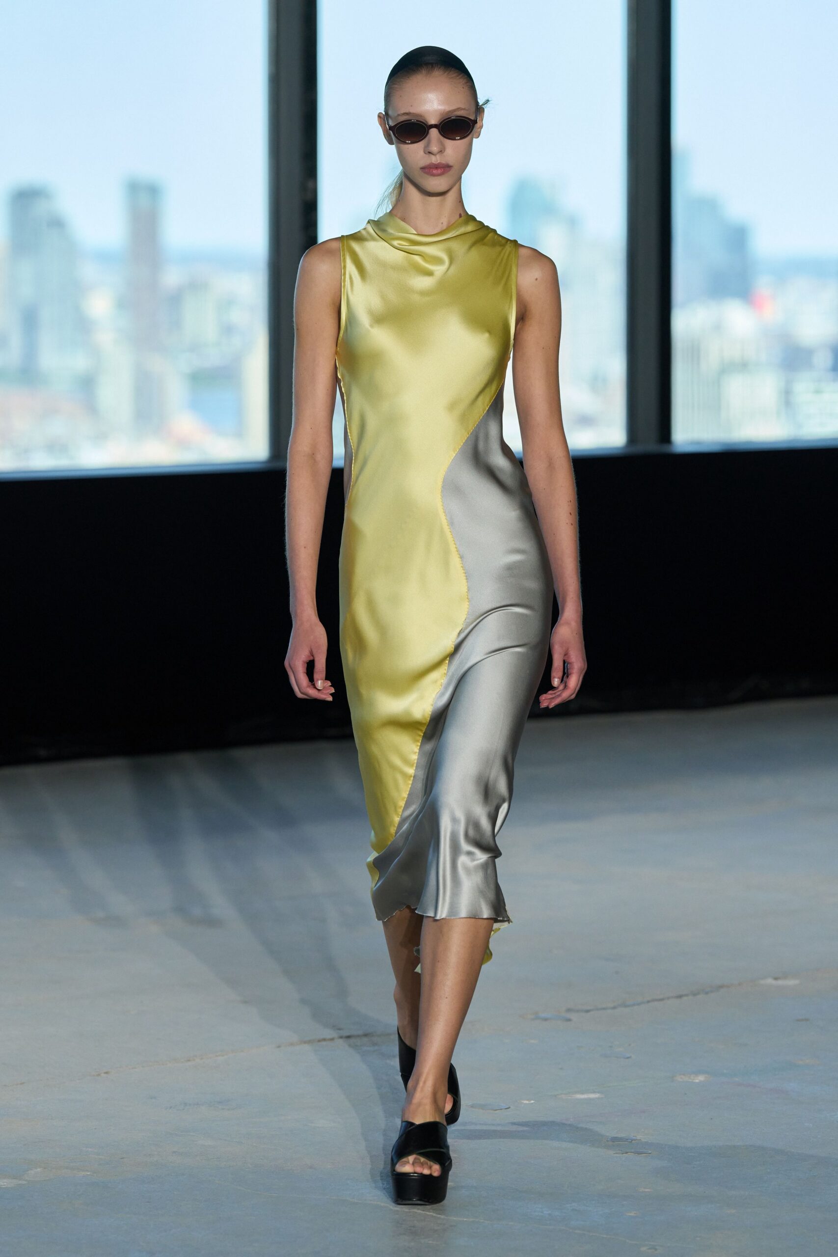 Narrow oval sunglasses with smoked lenses. Bias-cut satin slip dress in chartreuse and dove grey, joined by a snaking overlocked seam. Black platform slide mules. Crossing a hotel atrium early, before the conference badges come out.