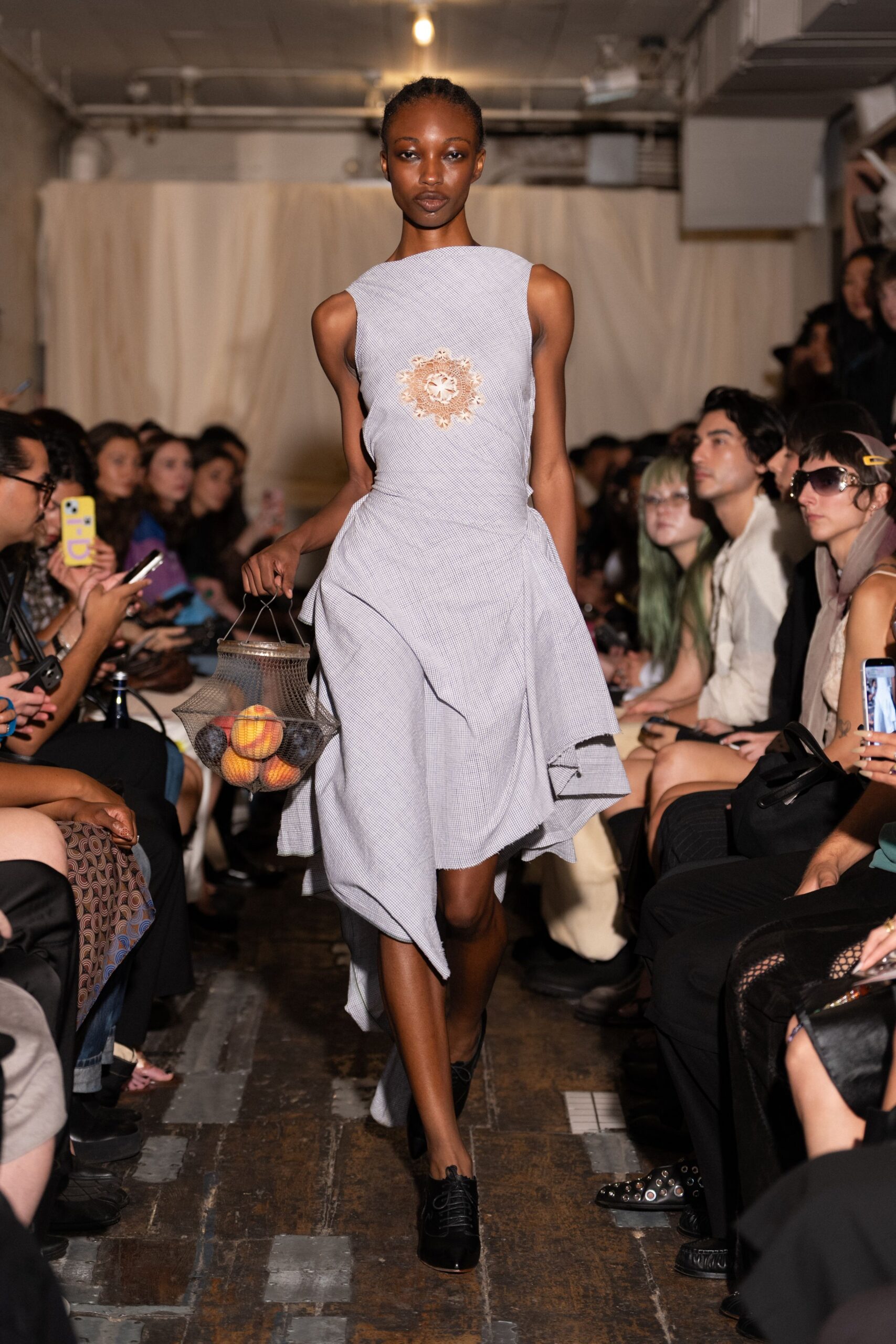 Pale micro-gingham dress with a bateau neck and raw uneven hem. Tea-stained crochet medallion centered on the chest. Wire mesh market basket filled with peaches and dark plums. Black lace-up heels with a cutaway instep. Walking back from the greengrocer before the fruit bruises in heat.