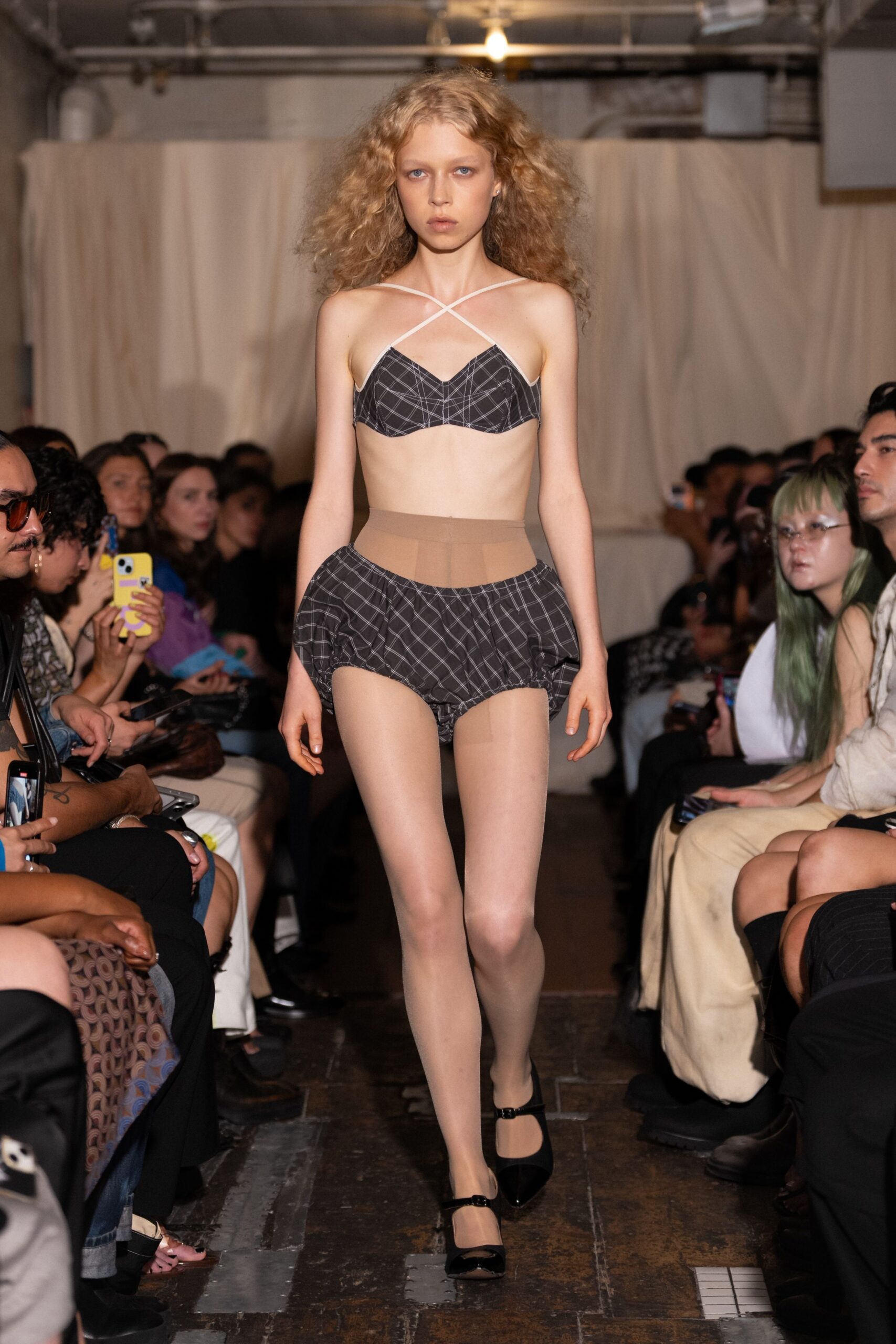 Charcoal plaid bralette with white straps crossing high on the chest. Matching bloomer briefs, softly gathered into rounded hips. Sheer nude tights with a darker control-top band. Black patent Mary Janes with a narrow instep strap. For buying bodega flowers still in yesterday's clothes.