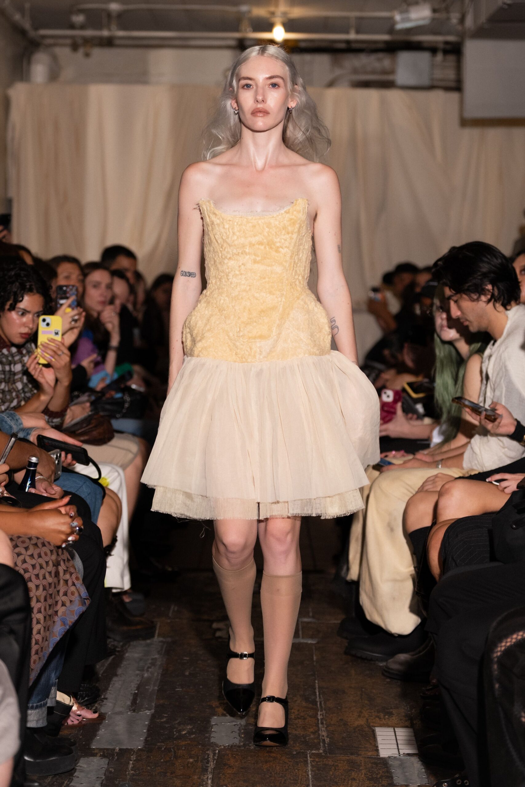 Butter-cream crushed velvet bodice, raw strapless edge above a stiff tulle skirt. Beige knee socks, pulled smooth. Black patent Mary Janes with narrow double straps. For cake cutting in a rented hall where the parquet sticks to your shoes.